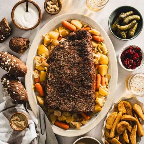 sliced cooked corned beef brisket on a serving platter with cooked veggies underneath and to the side, glasses with beer, bowls with pickled veggies, thick cut fries, bread rolls