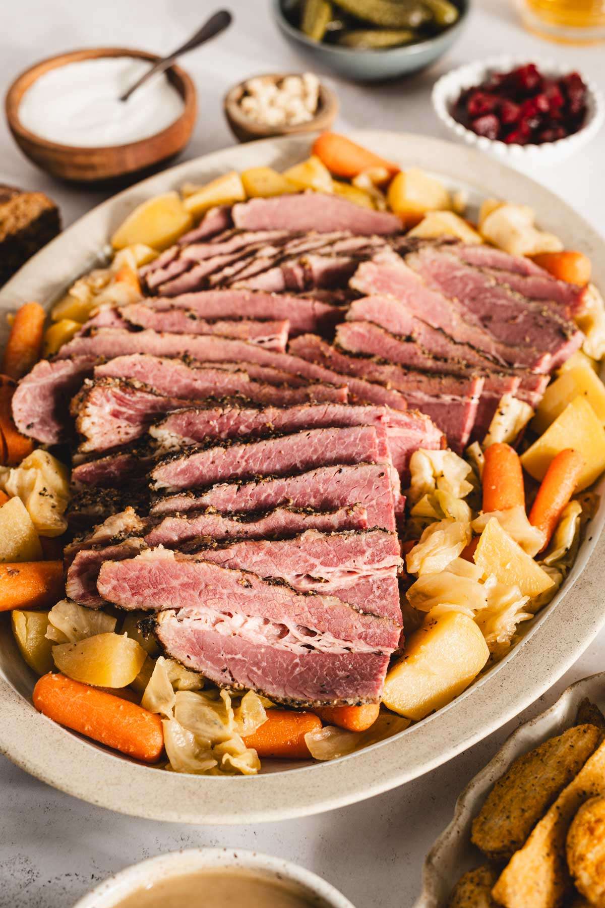 sliced cooked corned beef brisket on a serving platter with cooked veggies underneath and to the side, glasses with beer, bowls with pickled veggies, thick cut fries, bread rolls