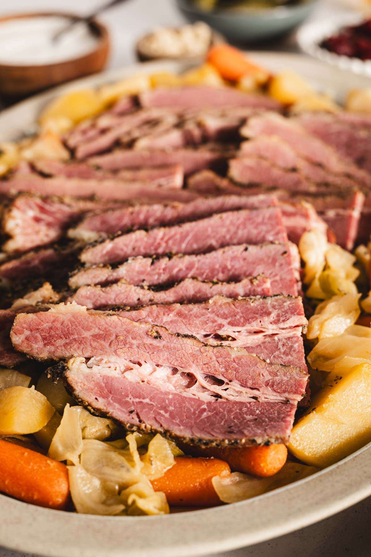 sliced cooked corned beef brisket on a serving platter with cooked veggies underneath and to the side, glasses with beer, bowls with pickled veggies, thick cut fries, bread rolls