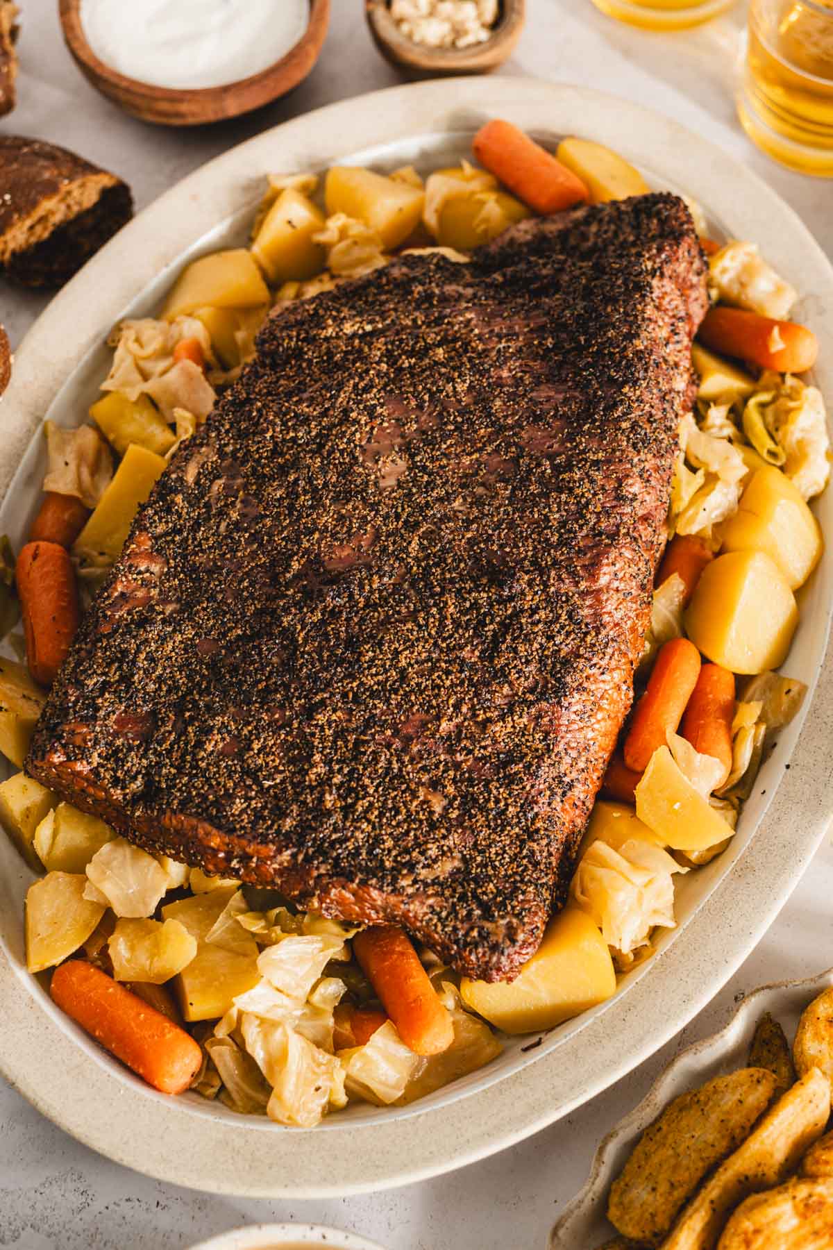 cooked corned beef brisket on a serving platter with cooked veggies underneath and to the side, glasses with beer, bowls with pickled veggies, thick cut fries, bread rolls