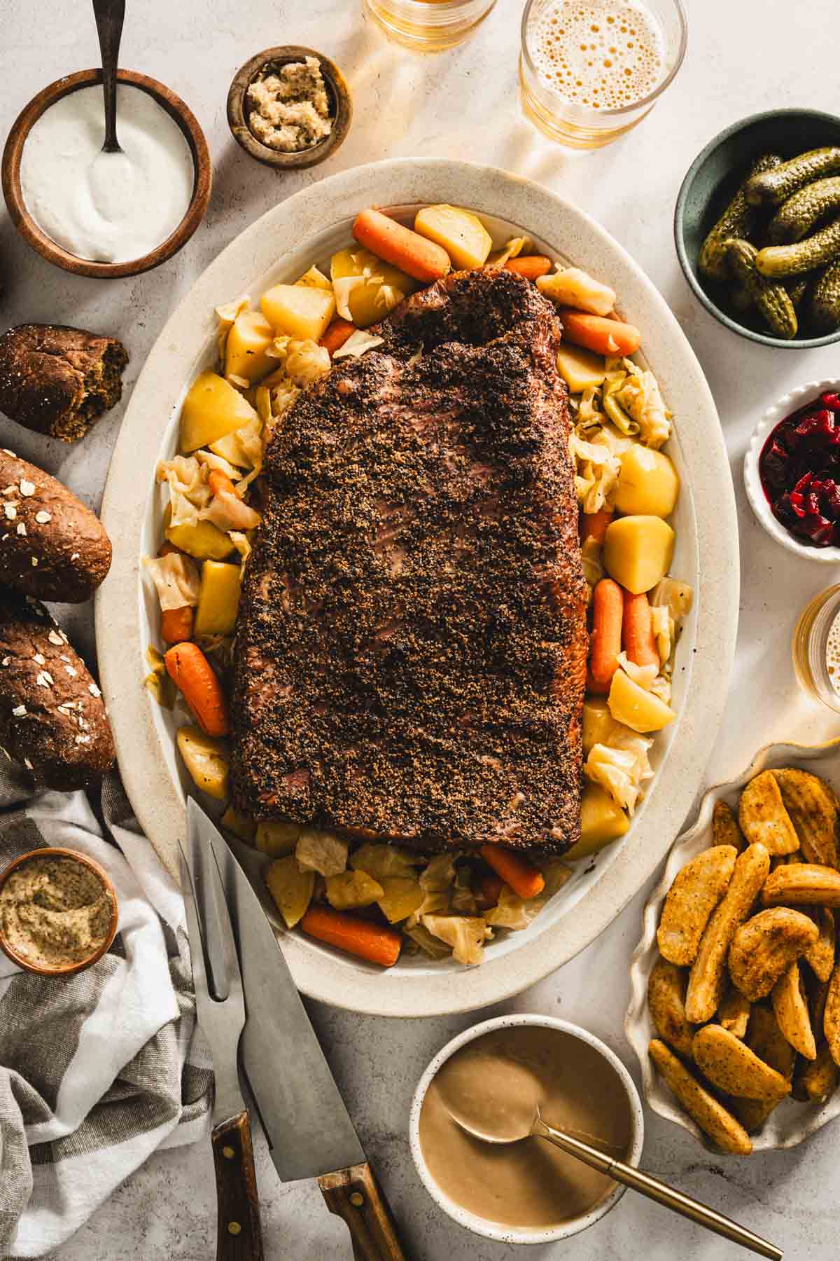 cooked corned beef brisket on a serving platter with cooked veggies underneath and to the side, glasses with beer, bowls with pickled veggies, thick cut fries, bread rolls