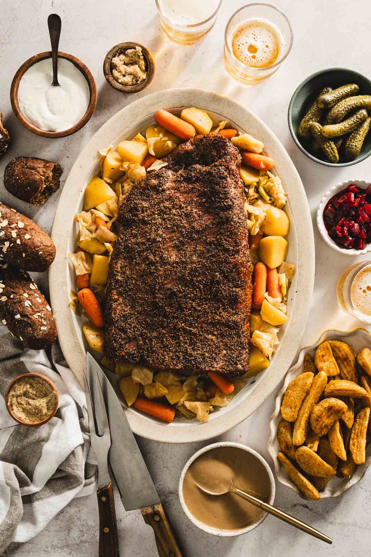 cooked corned beef brisket on a serving platter with cooked veggies underneath and to the side, glasses with beer, bowls with pickled veggies, thick cut fries, bread rolls