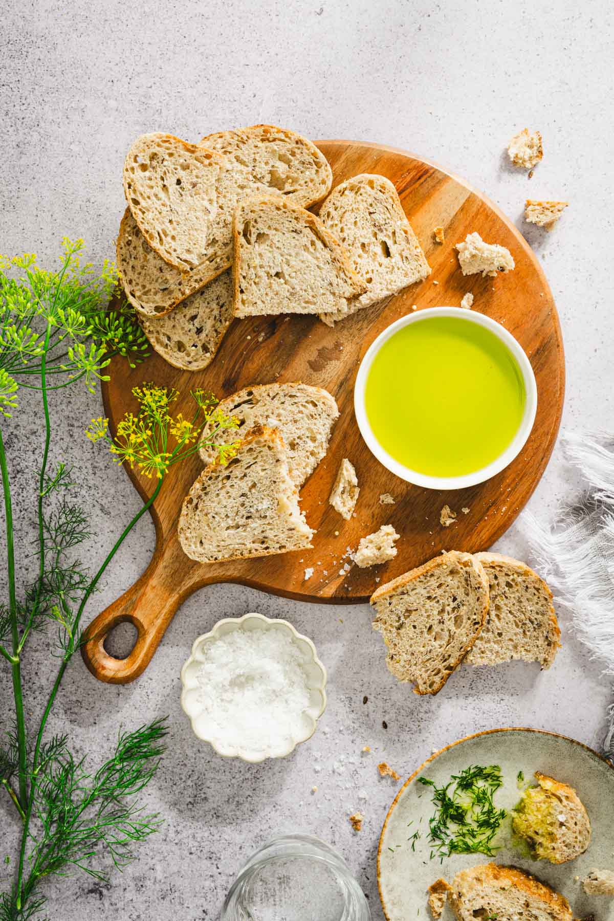 round wooden board with slices of bread, dish with dill oil, salt; few branches of fresh dill