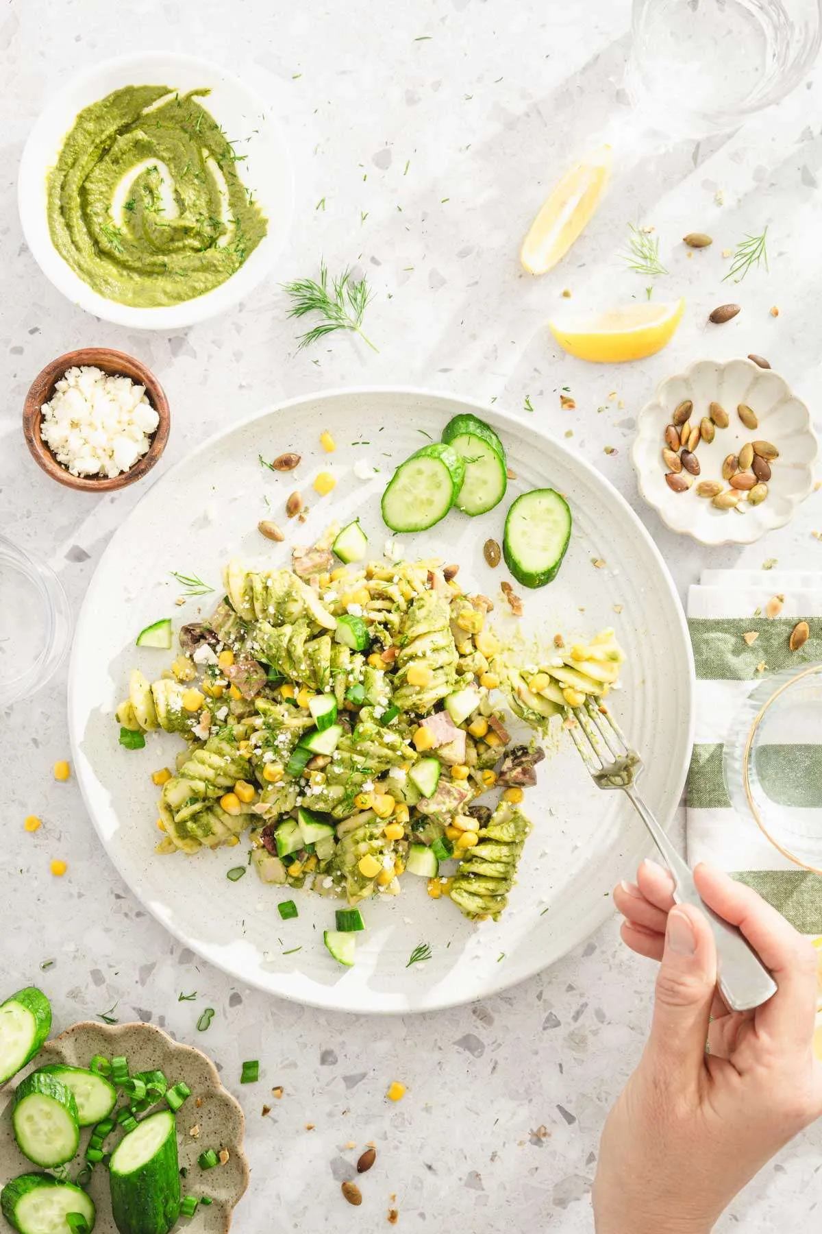 pesto pasta served on a plate, slices cucumbers, pumpkin seeds, hand in frame holding fork, dish with feta cheese