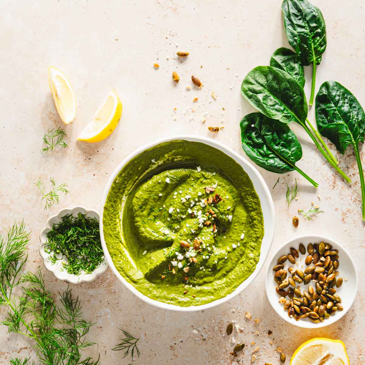 top view of garnished bowl of dill pesto, spinach leaves, dish with fresh dill and roasted pumpkin seeds, lemon wedges