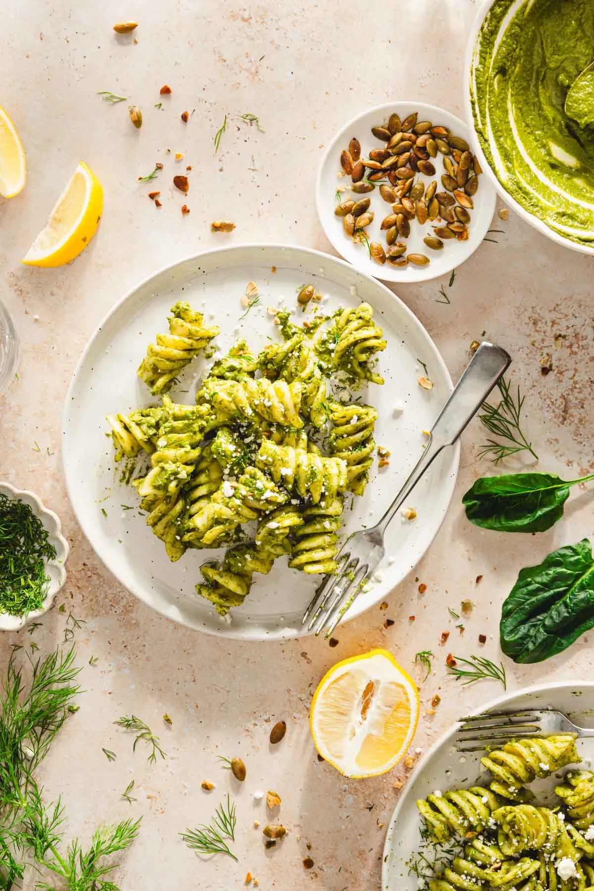 pasta mixed with pesto on a plate, spinach leaves, roasted seeds, lemon wedges, bowl of pesto