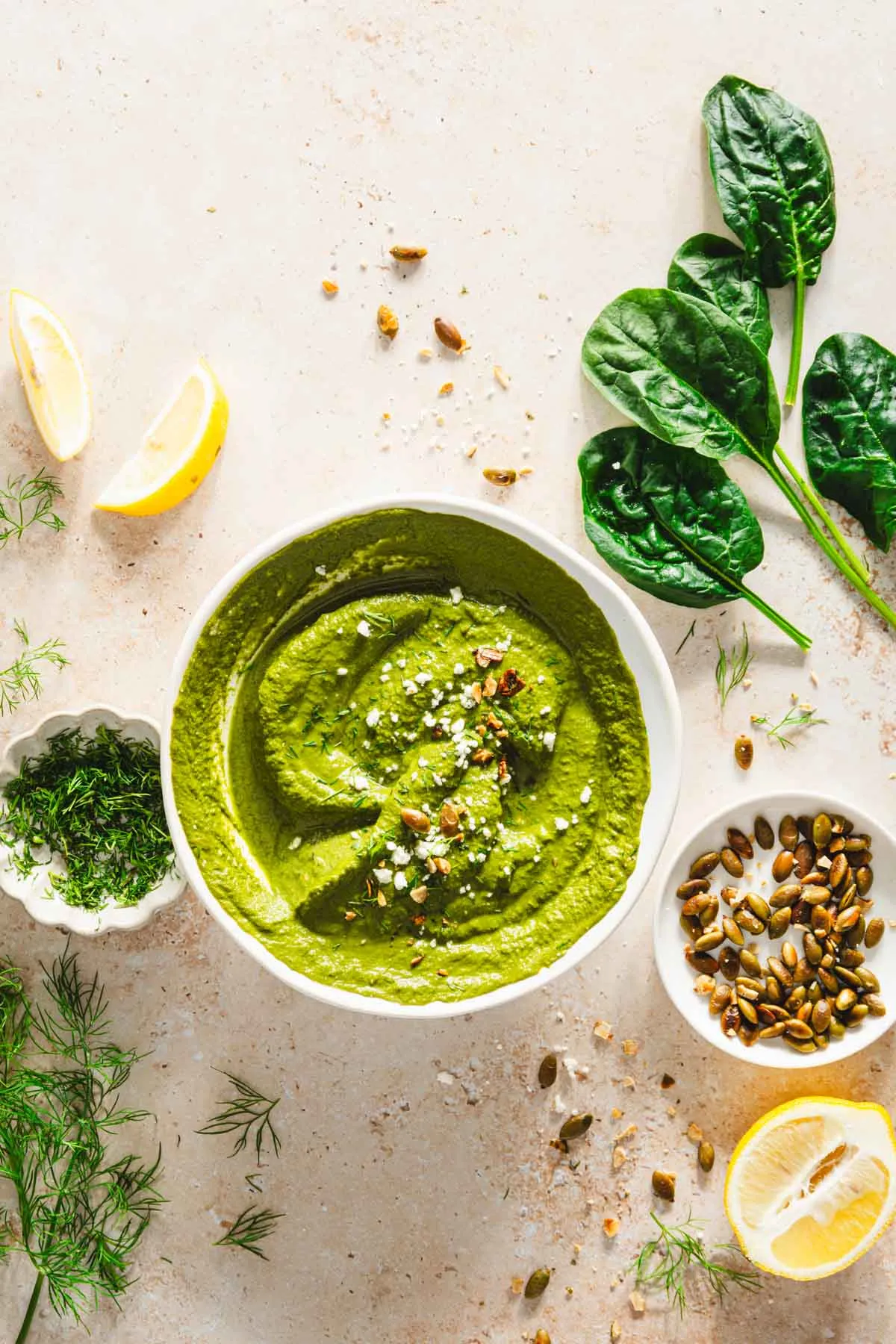 top view of garnished bowl of dill pesto, spinach leaves, dish with fresh dill and roasted pumpkin seeds, lemon wedges