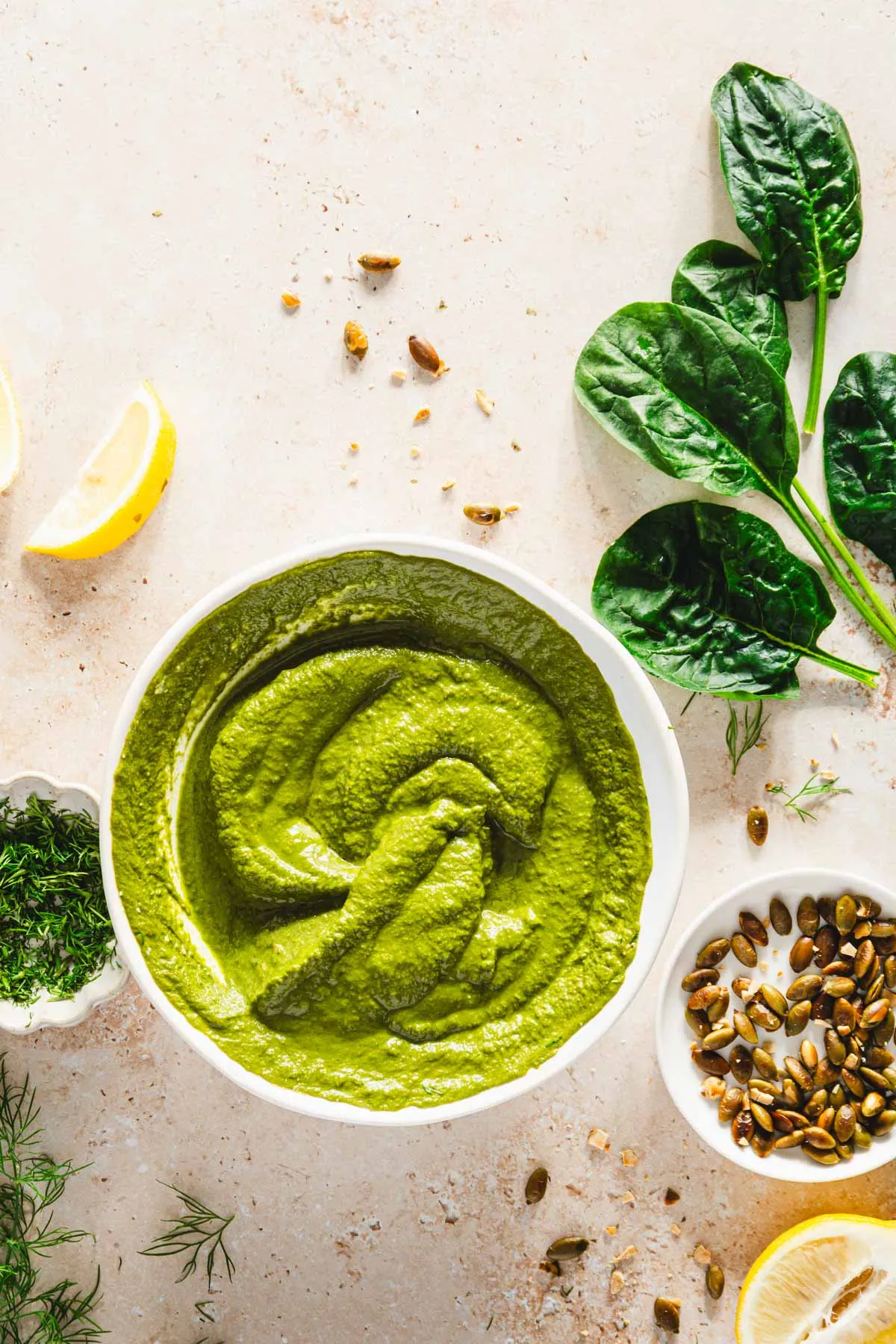 top view of plain bowl of dill pesto, spinach leaves, dish with fresh dill and roasted pumpkin seeds, lemon wedges