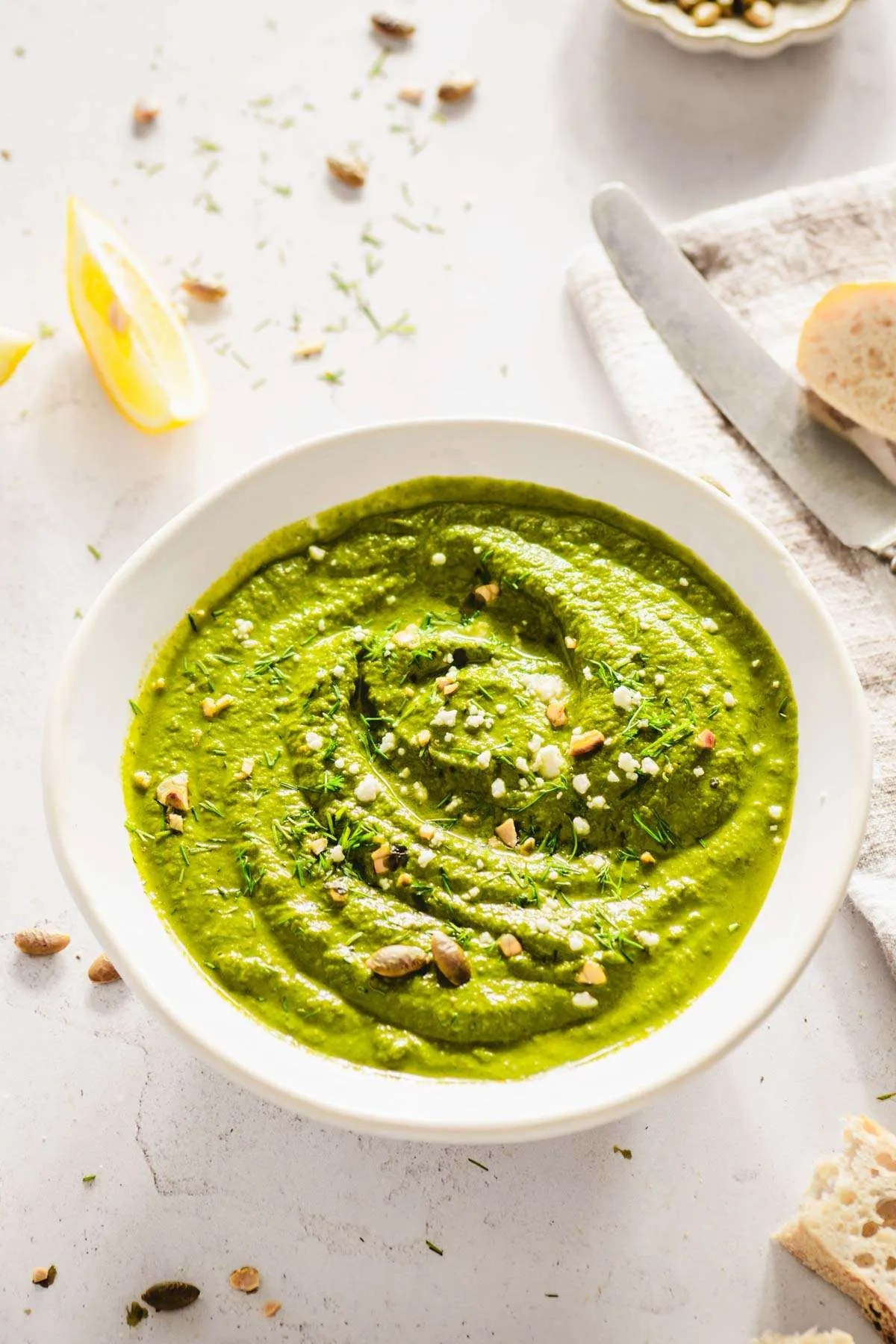 bowl with dill pesto garnished with crushed pumpkin seeds, chopped fresh dill, lemon wedges, bread slices, butter knife, dish with pumpkin seeds