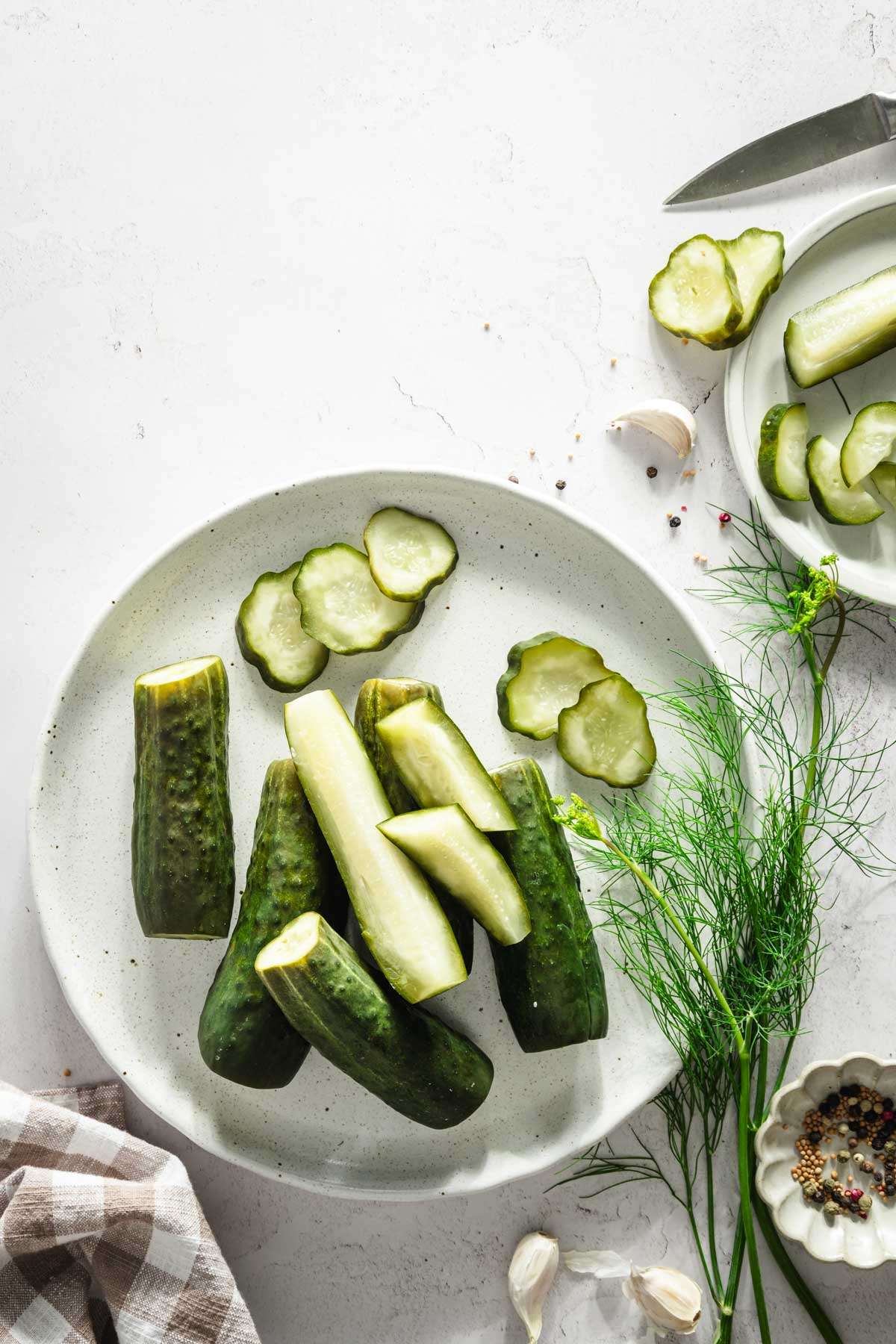 half sour pickled arranged on a plate, some cut in half, some sliced, fresh dill, dish with spices, small knife