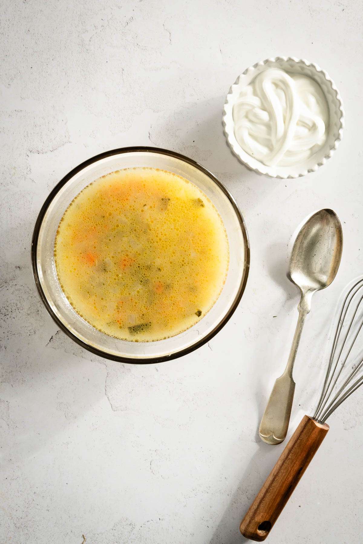 small glass bowl with the dill pickle soup, small dish with sour cream, spoon and whisk on the side