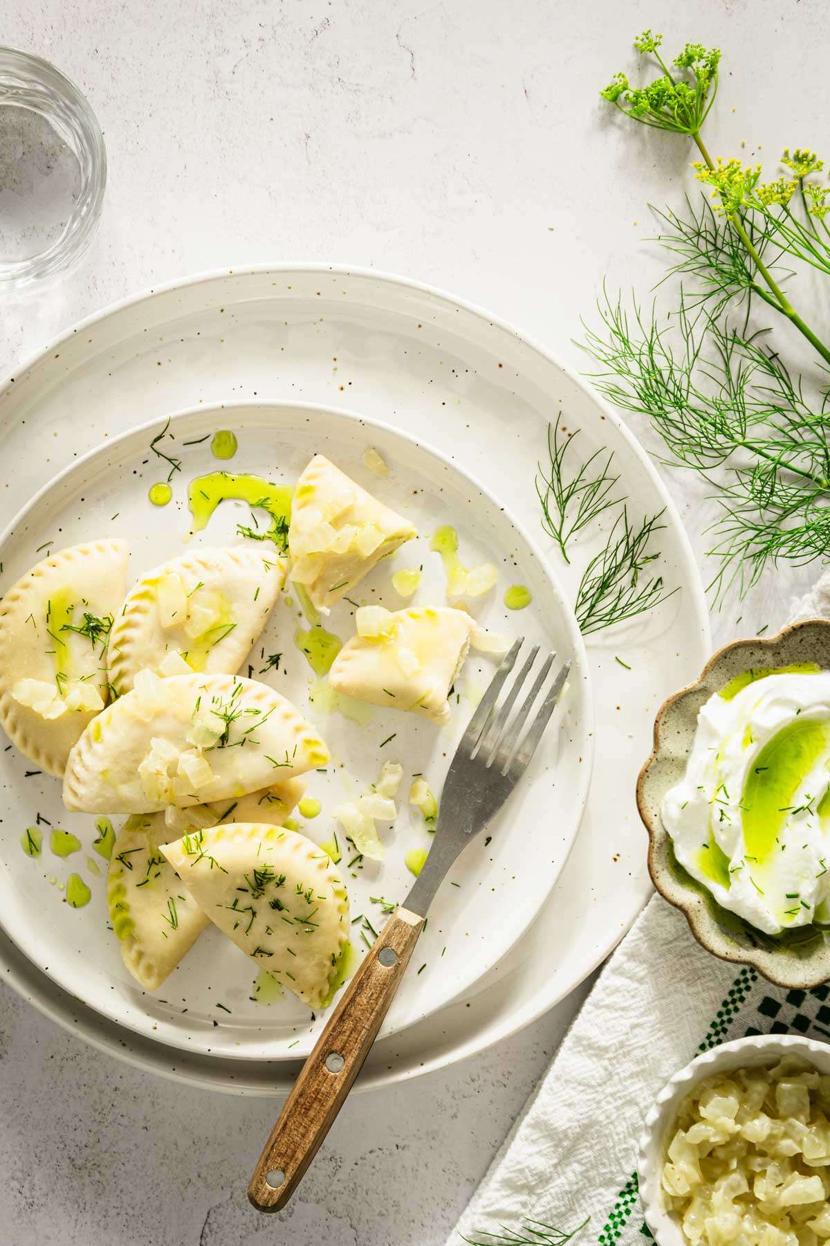 dill pierogi on a plate, garnished with fresh dill, onion and oil, fork and glass with water, dish with cooked onions and one with yogurt and oil