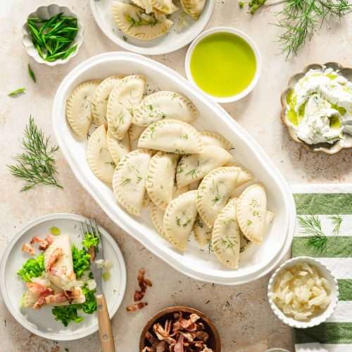 dill pierogis arranged on a oval platter, two plates with pierogis, dish with green onions, fried bacon, cooked onions and dill oil