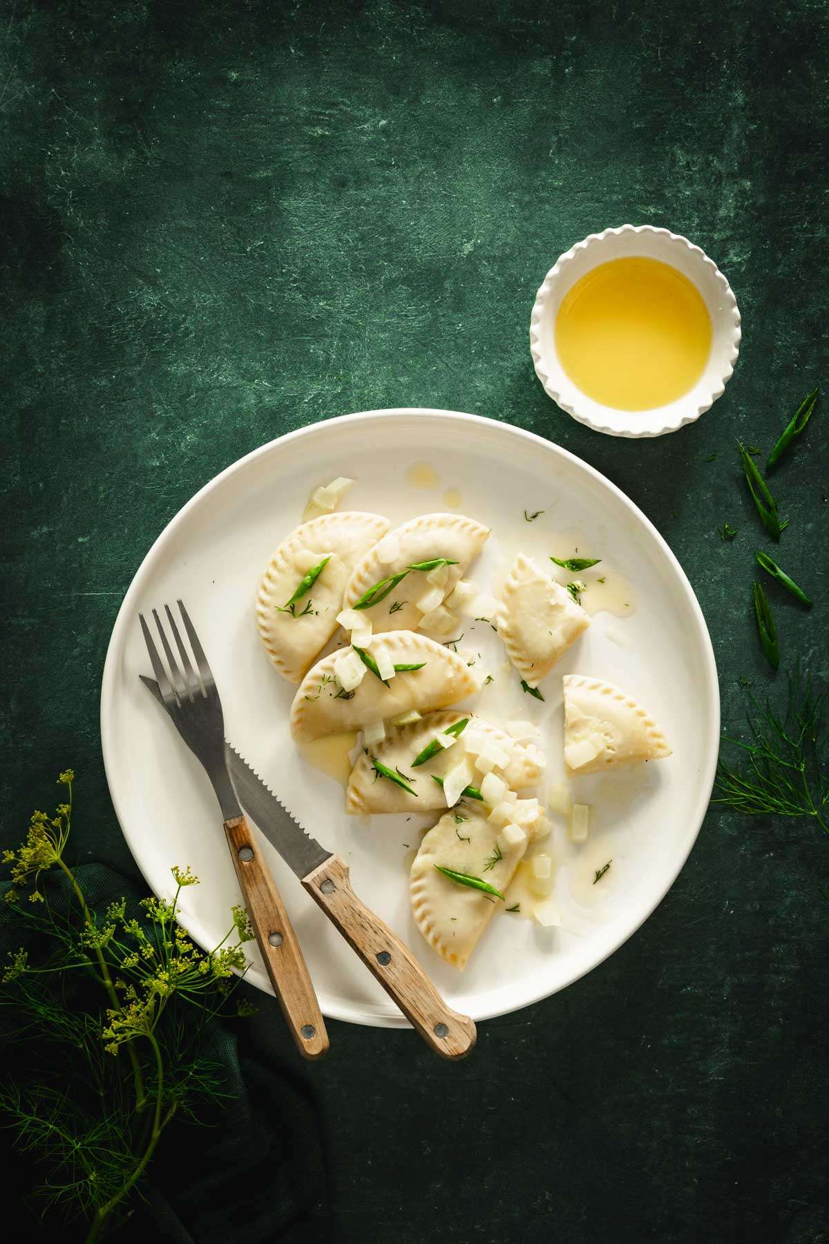 plate with dill pierogis on a dark green background, dish with melted butter, few branches of fresh dill