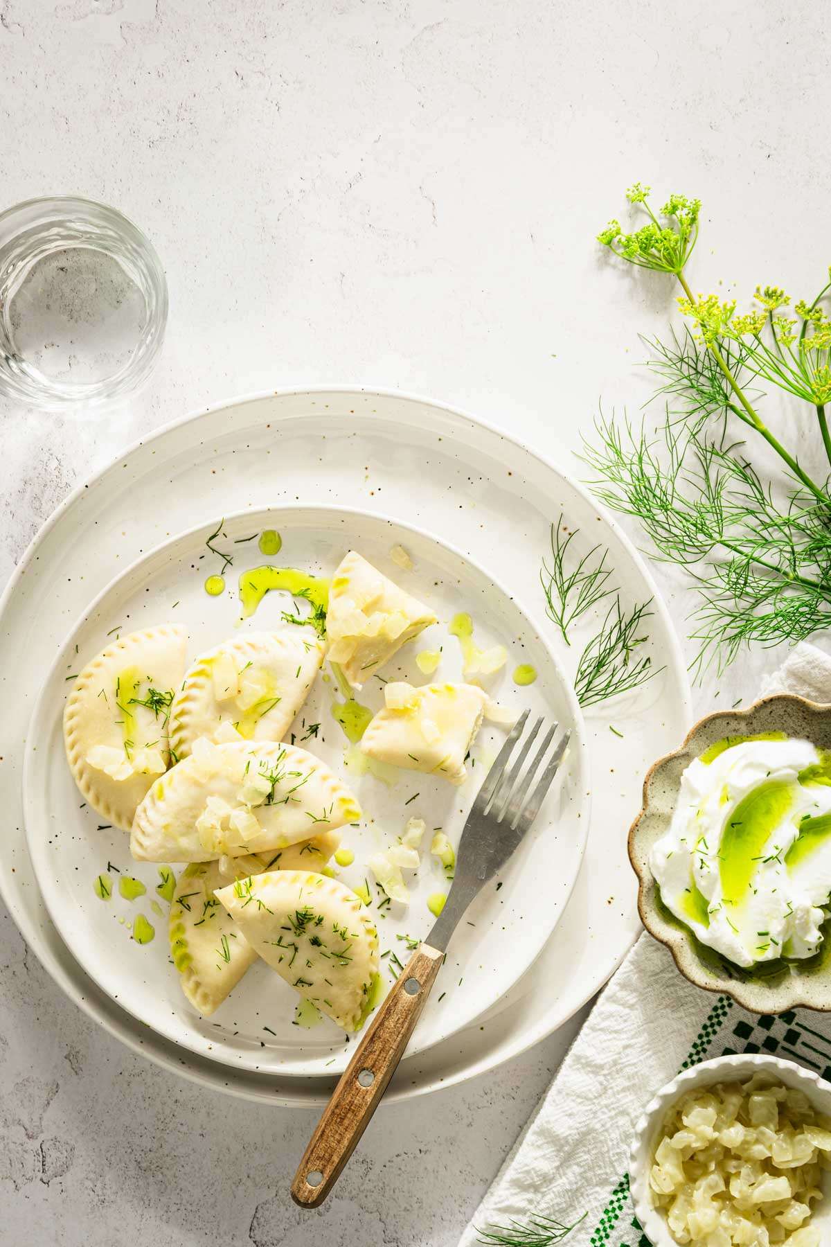 dill pierogi on a plate, garnished with fresh dill, onion and oil, fork and glass with water, dish with cooked onions and yogurt, dill branches