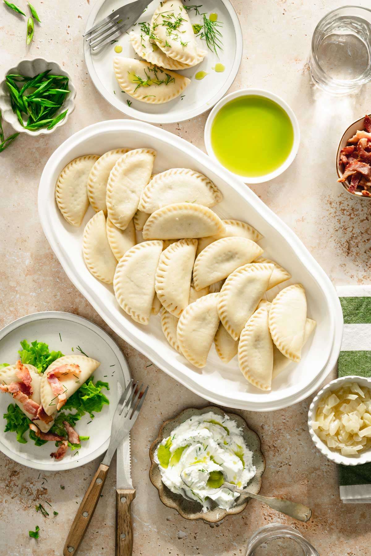 dill pierogis arranged on a oval platter, two plates with pierogis, dish with green onions, fried bacon, cooked onions and dill oil