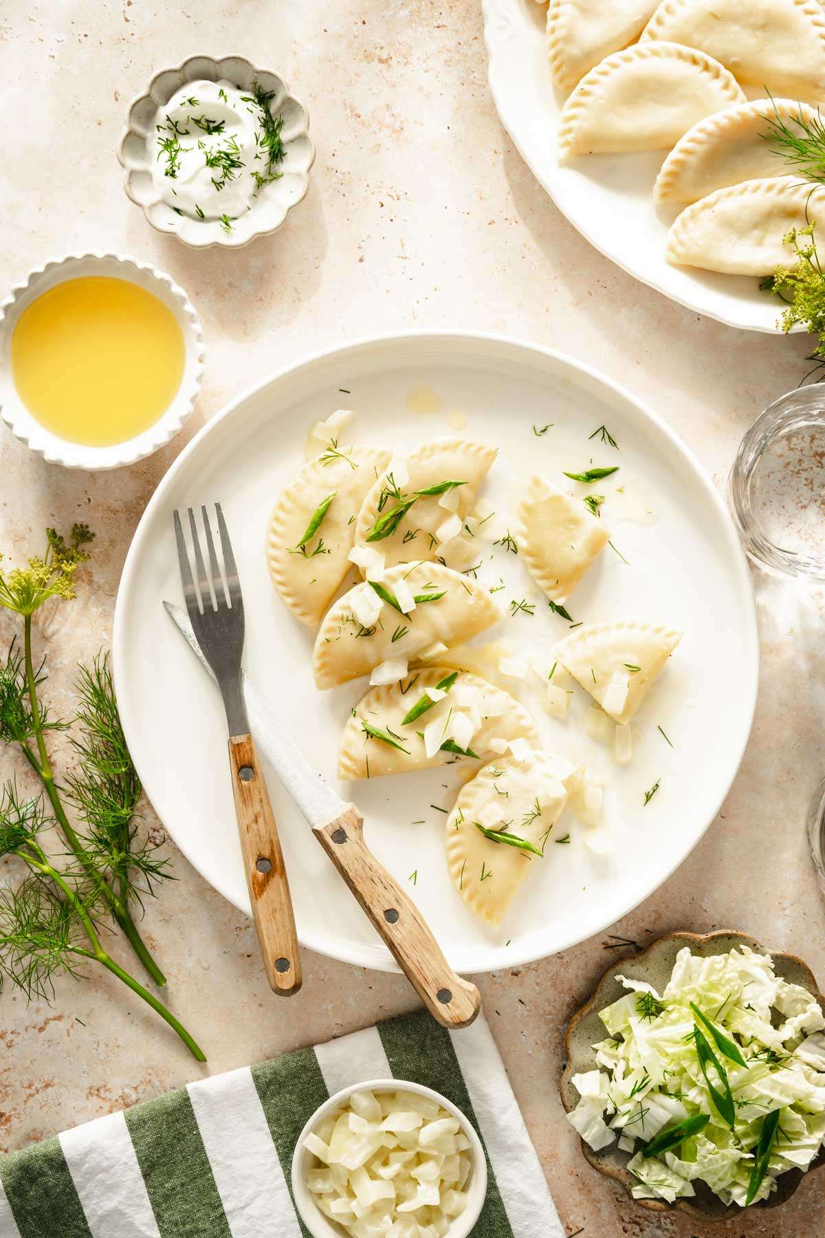 plate with pierogis garnished with fried onion and chives, side salad, dish with cooked onions, platter with cooked pierogis, dish with melted butter and yogurt