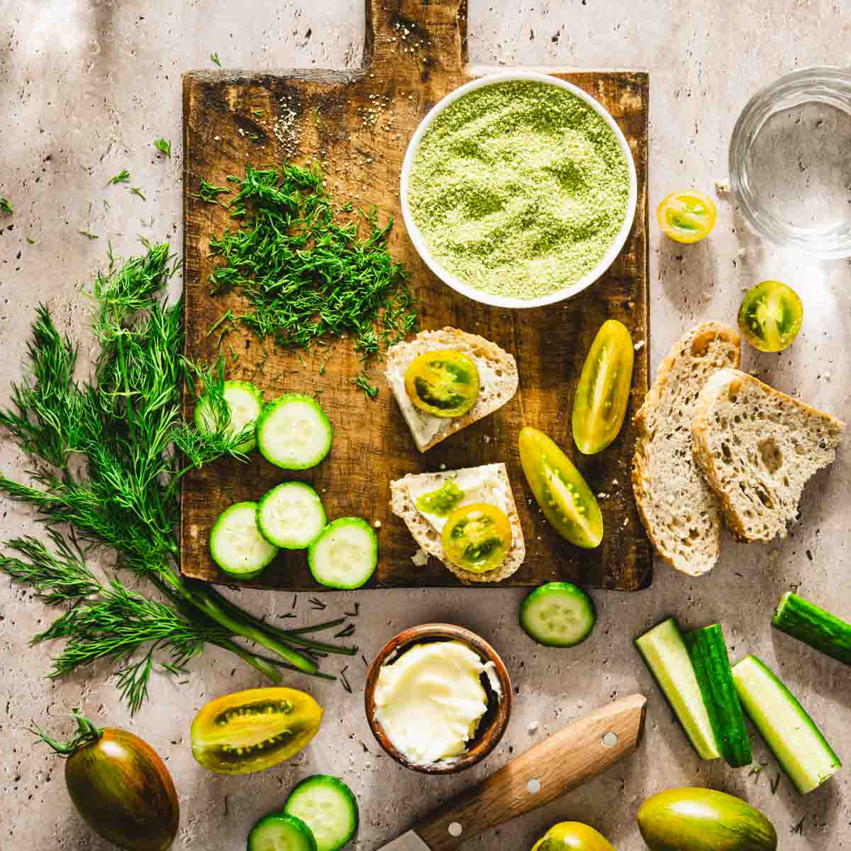 cutting board with dish with dill salt, cut up tomatoes, cucumbers, chopped dill, few slices of bread, dish with butter, glasses with water