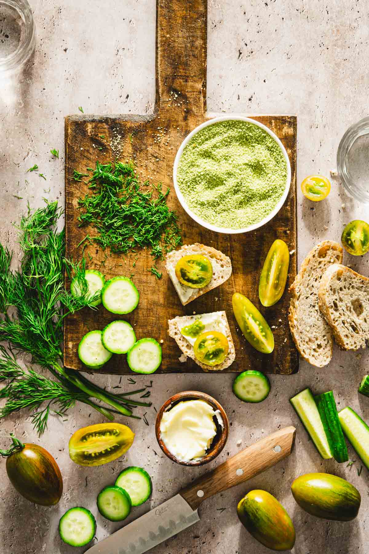 cutting board with dish with dill salt, cut up tomatoes, cucumbers, chopped dill, few slices of bread, dish with butter, glasses with water