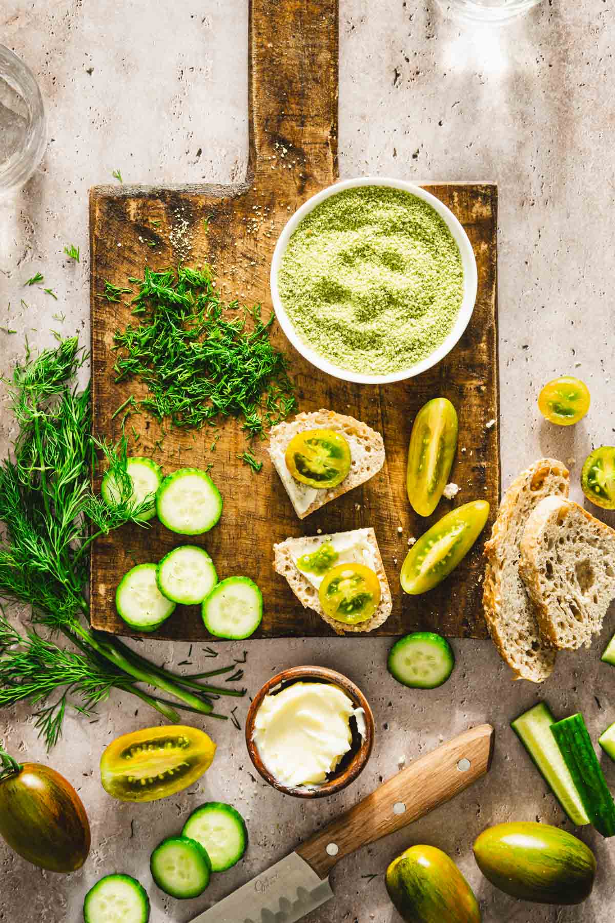 cutting board with dish with dill salt, cut up tomatoes, cucumbers, chopped dill, few slices of bread, dish with butter, glasses with water