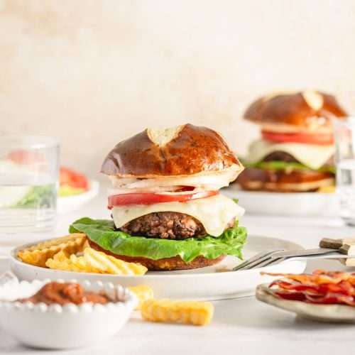 two elk burgers served on a pretzel bun, presented on plates, dish with ketchup, glass of water, fries