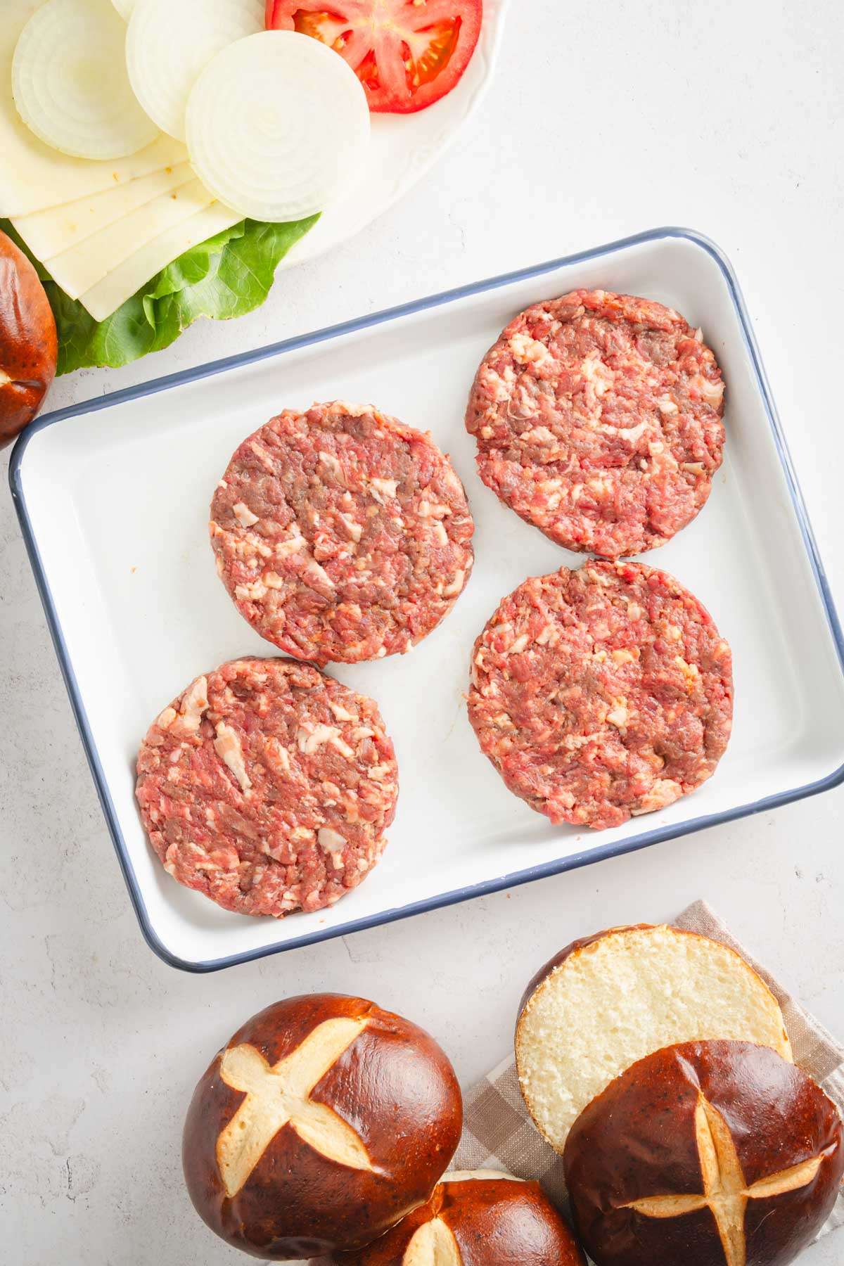 raw, shaped elk burgers on a metal tray, pretzel buns, dish with lettuce, tomato, onion and cheese