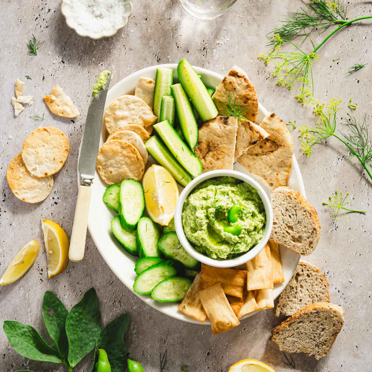 platter with veggies, bread and crackers and a bowl with fava bean hummus; lemon slices, glass of water, bread