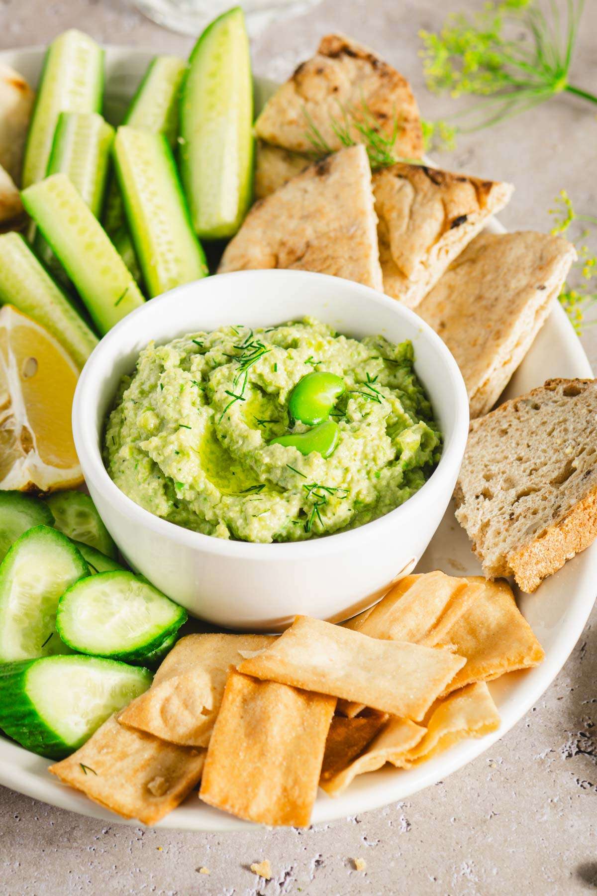 close up of platter with veggies, bread and crackers and a bowl with fava bean hummus; lemon slices, glass of water, bread