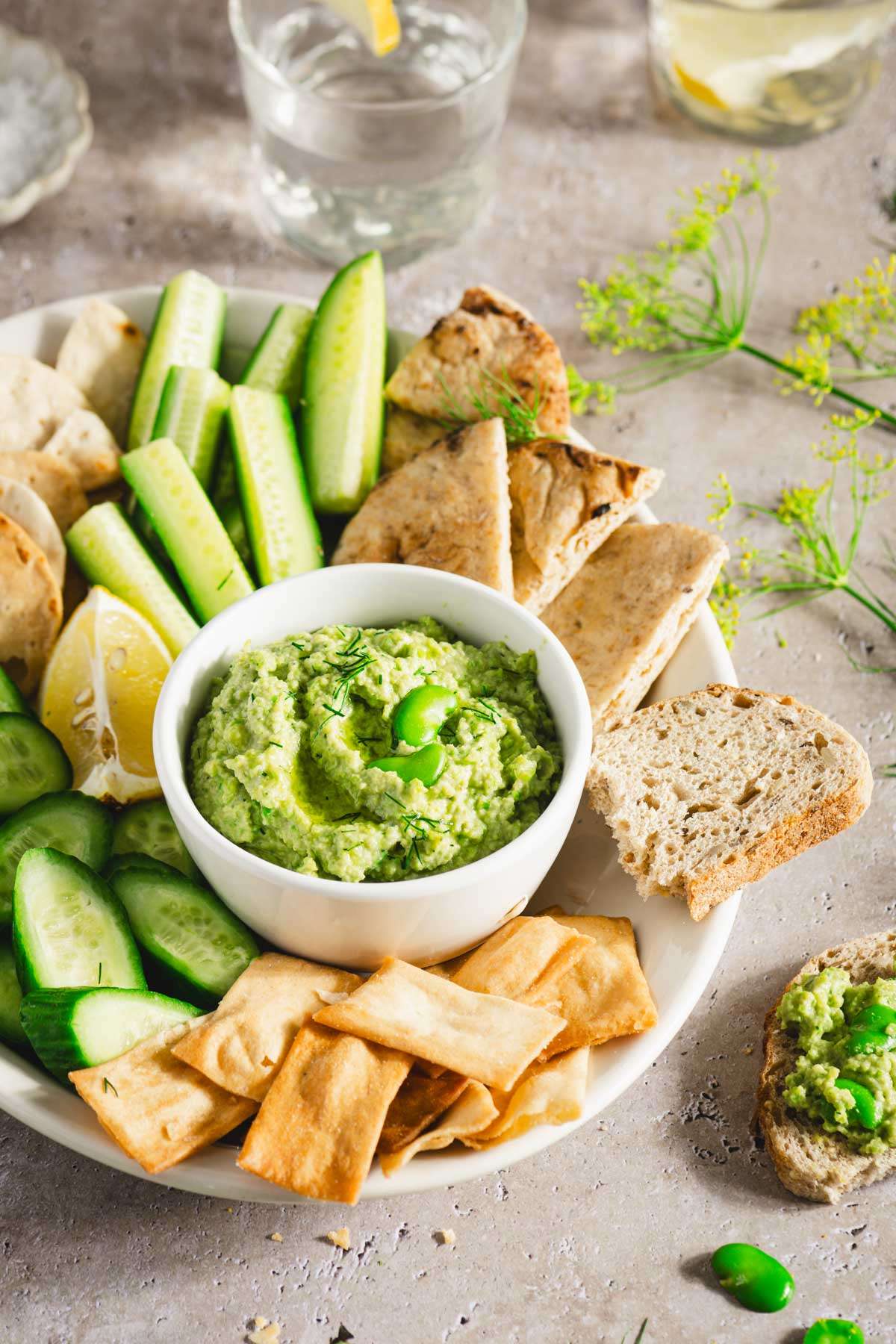 platter with veggies, bread and crackers and a bowl with fava bean hummus; lemon slices, glass of water, bread