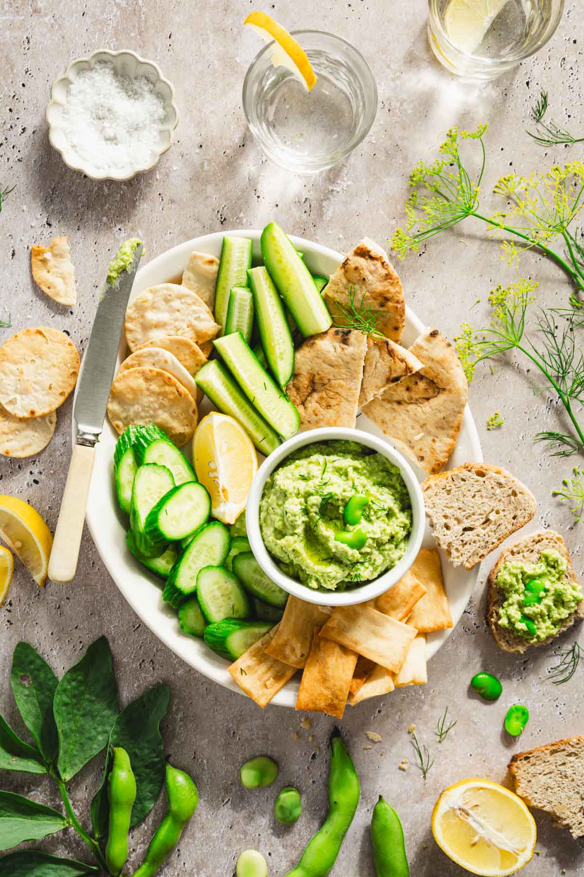 platter with veggies, bread and crackers and a bowl with fava bean hummus; lemon slices, glass of water, bread