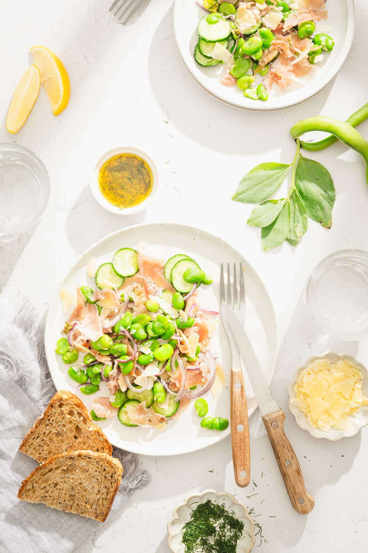 fava bean salad in a serving bowl, dish with fresh dill, parm cheese, slices of bread, glass with water