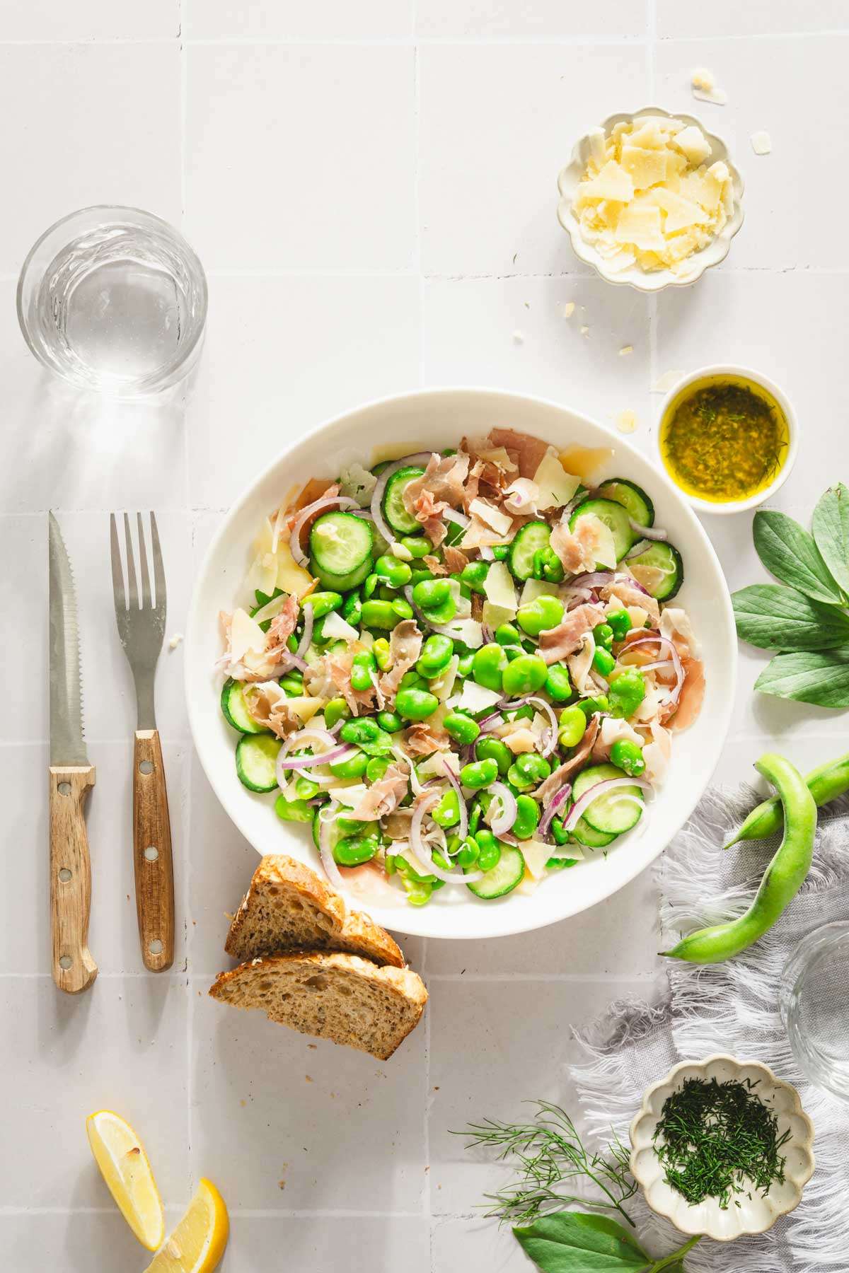 fava bean salad in a serving bowl, glass with water, dish with dressing, parm cheese, slices of bread