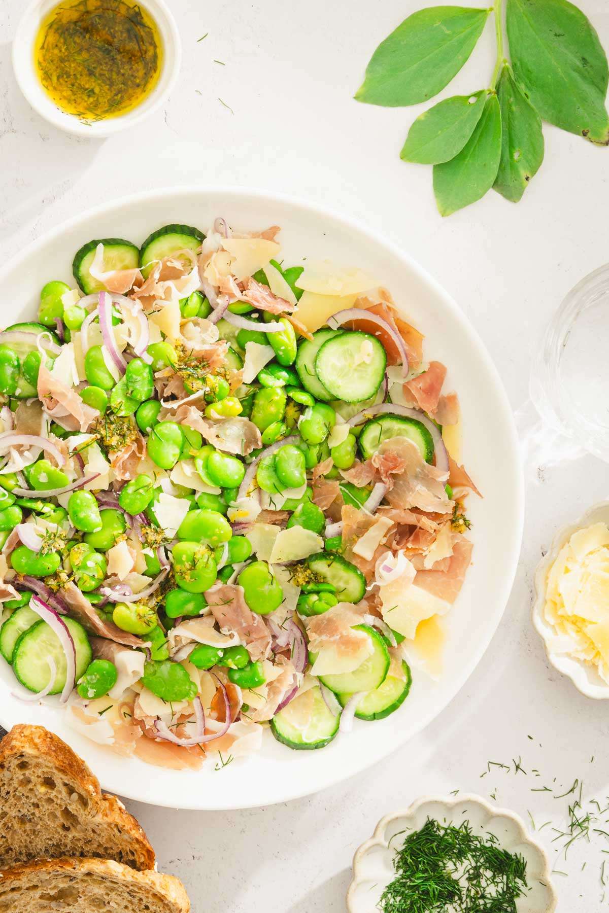 fava bean salad in a serving bowl, dish with fresh dill, parm cheese, slices of bread, glass with water