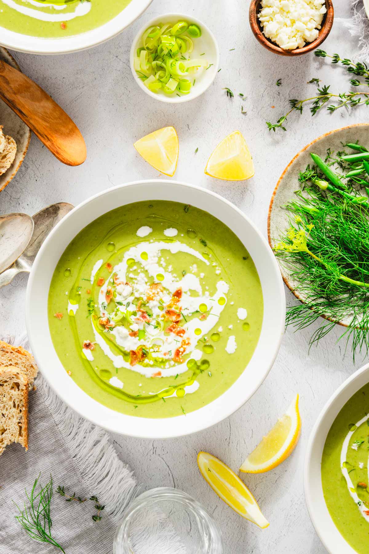 bowl with fava bean soup, garnished with cream, feta cheese and pieces of prosciutto; lemon wedges, plate with fresh dill and chives