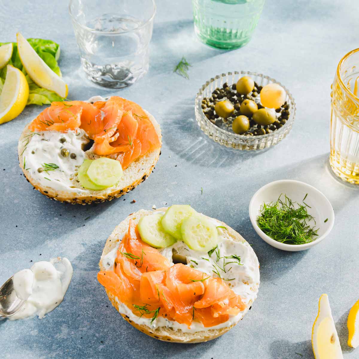 pieces of salmon on a bagel with cream cheese and sliced cucumbers, dish with olives, fresh dill; glasses with water