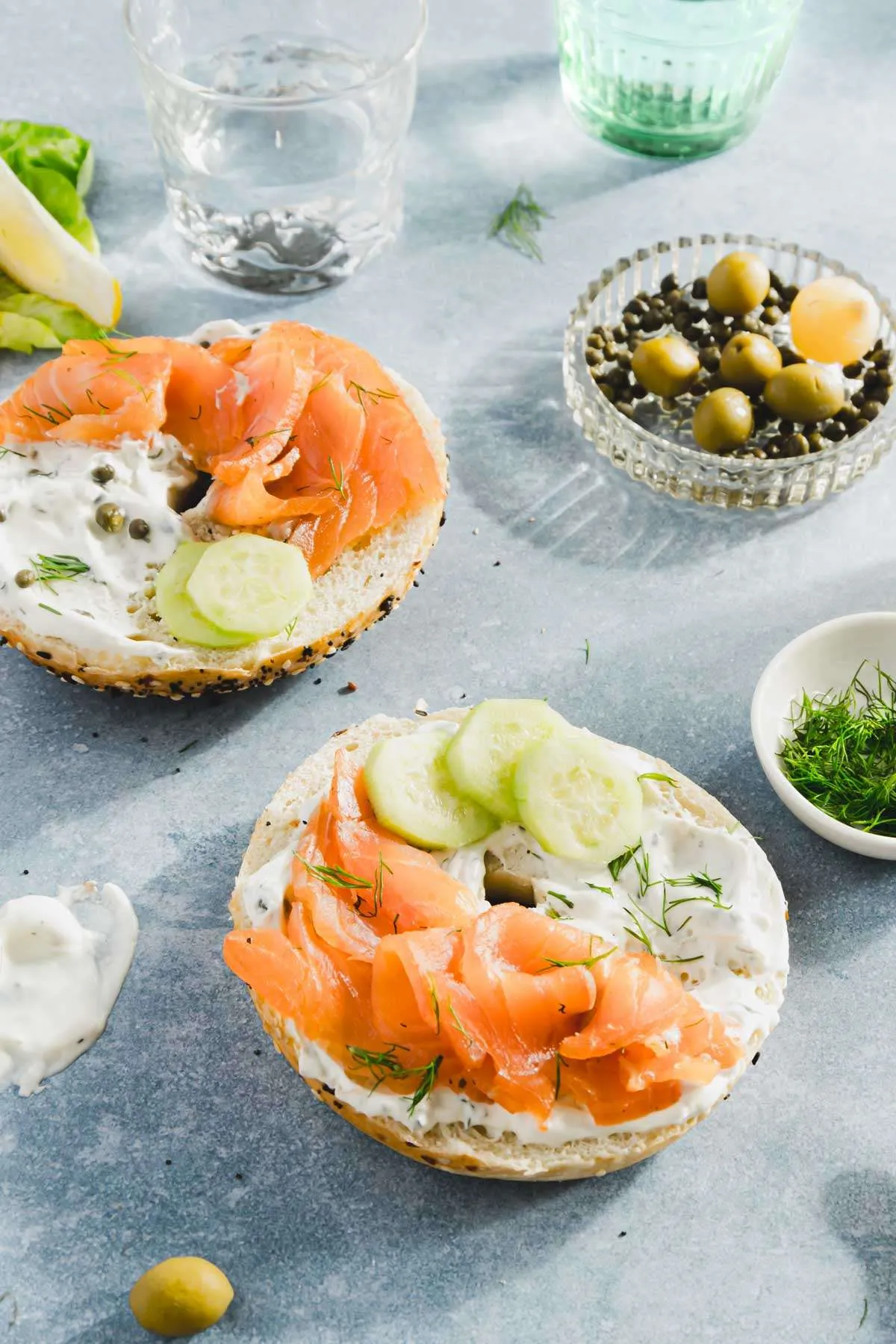pieces of salmon on a bagel with cream cheese and sliced cucumbers, dish with olives, fresh dill; glasses with water