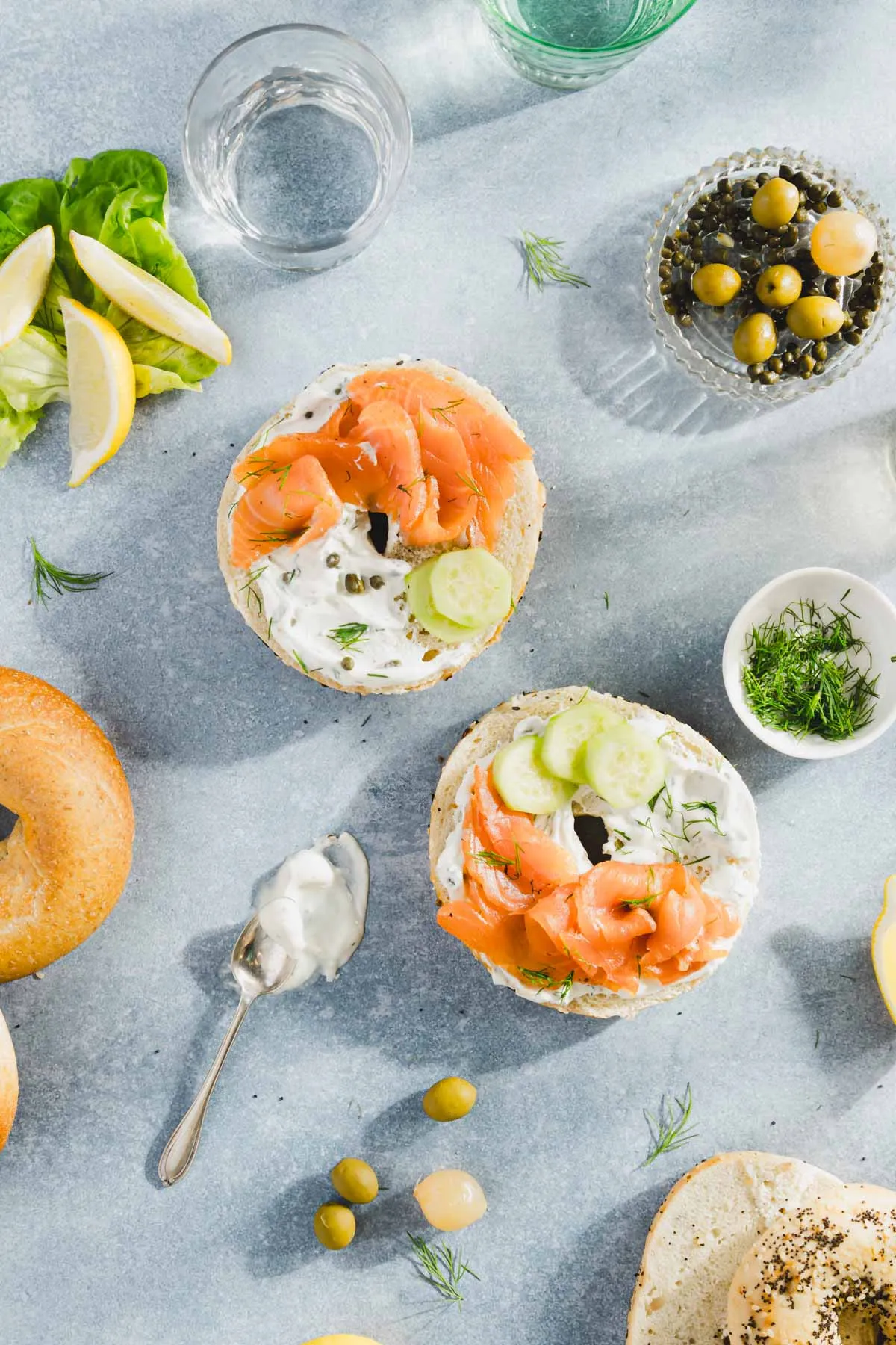 pieces of salmon on a bagel with cream cheese and sliced cucumbers, dish with olives, fresh dill; glasses with water