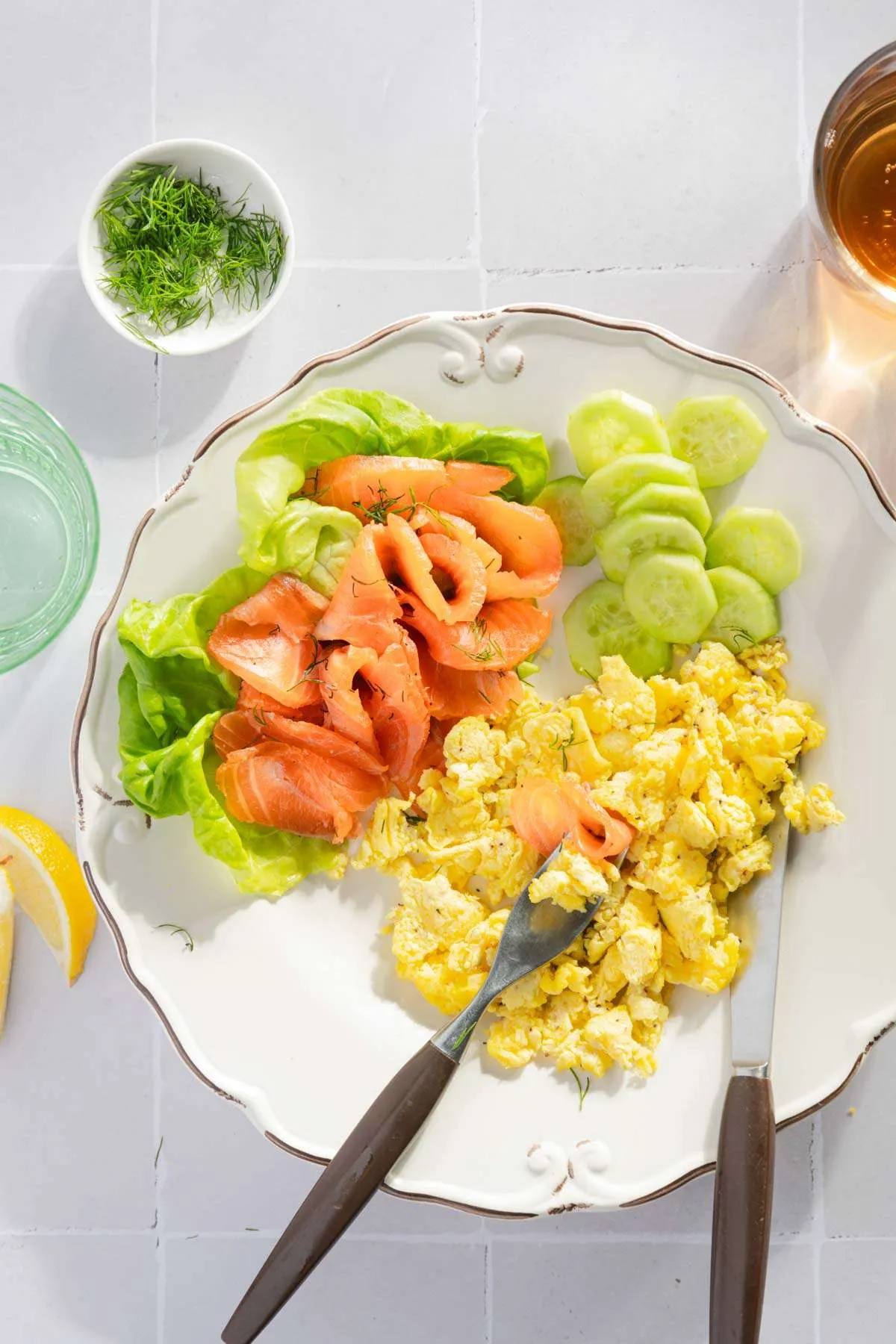 pieces of cured salmon on a plate next to scrambled eggs, lettuce leaf, lemon wedges, glass with water and tea, dish with fresh dill, sliced cucumber on the plate