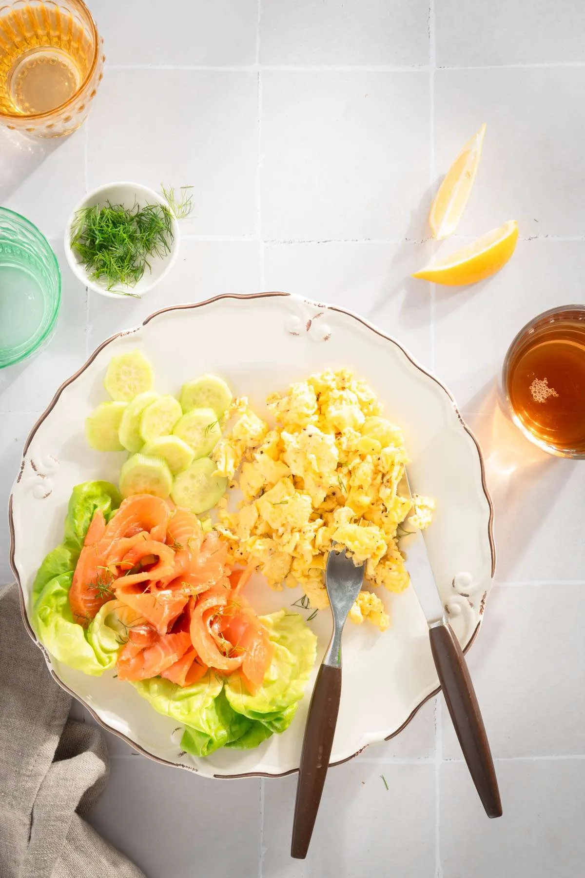 pieces of cured salmon on a plate next to scrambled eggs, lettuce leaf, lemon wedges, glass with water and tea, dish with fresh dill