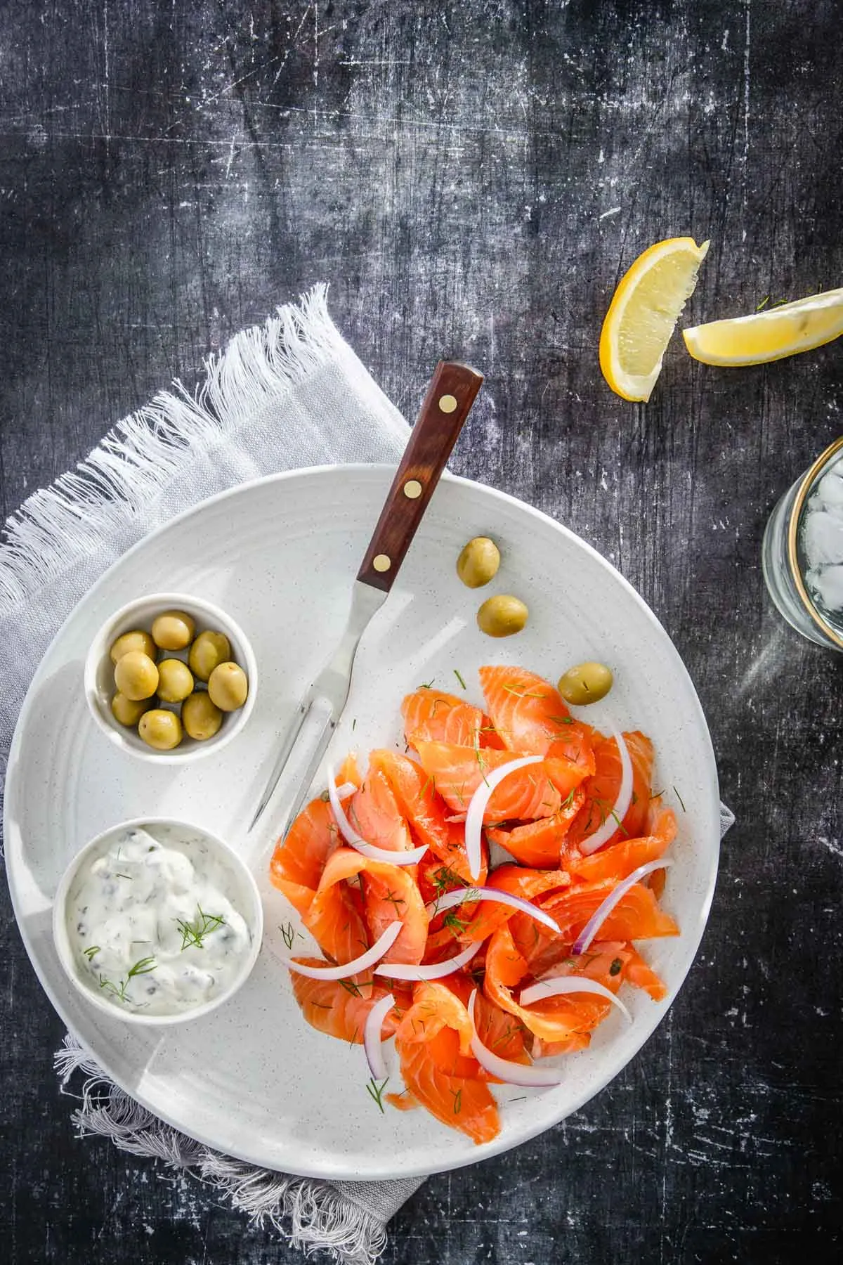 slices of cured salmon on serving plate, with olives and yogurt sauce, lemon wedges, glass with water