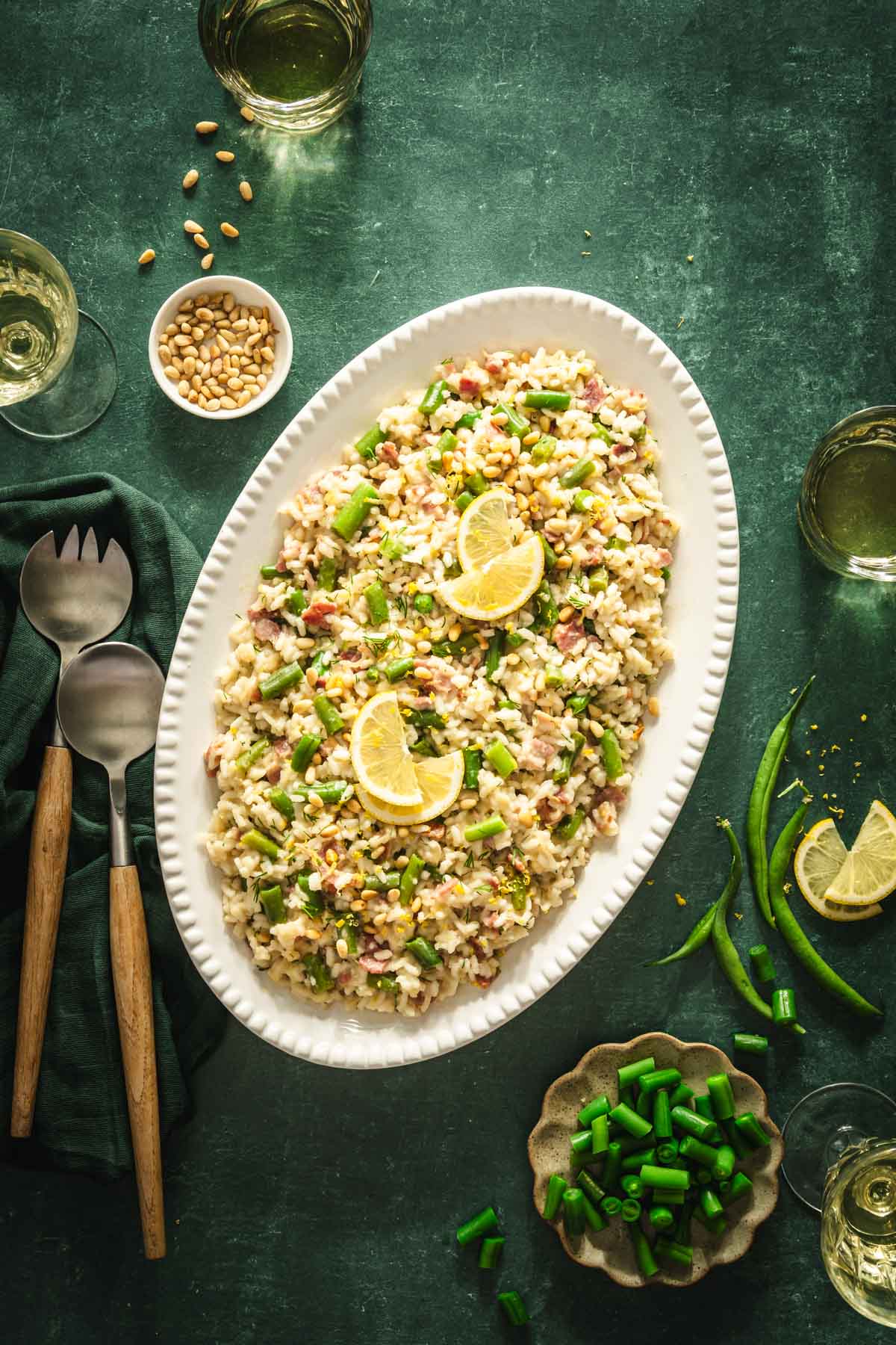 green background, risotto served in an oval plate, utensils, dish with pine nuts, glasses with wine, dish with green beans