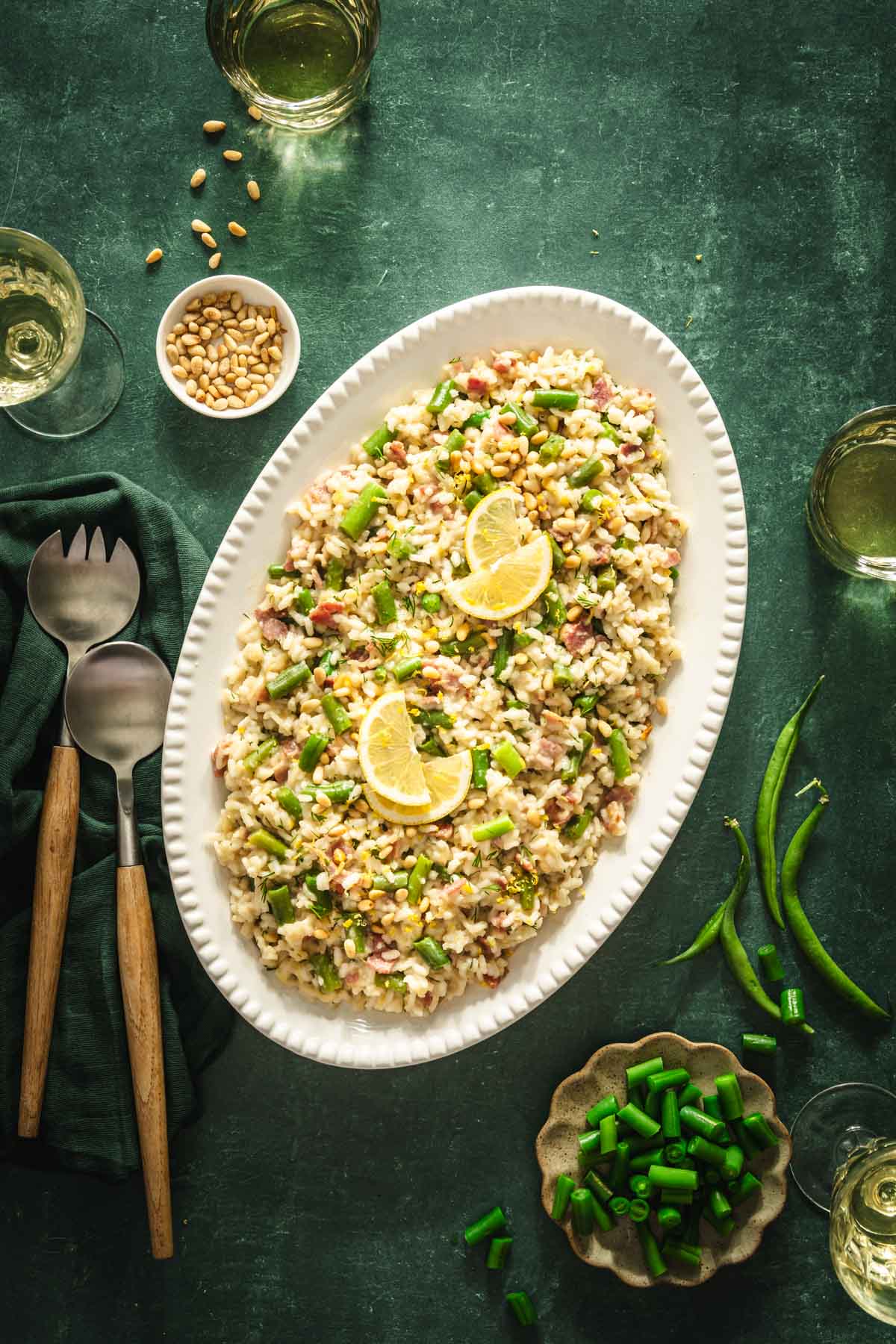 green background, risotto served in an oval plate, utensils, dish with pine nuts, glasses with wine, dish with green beans