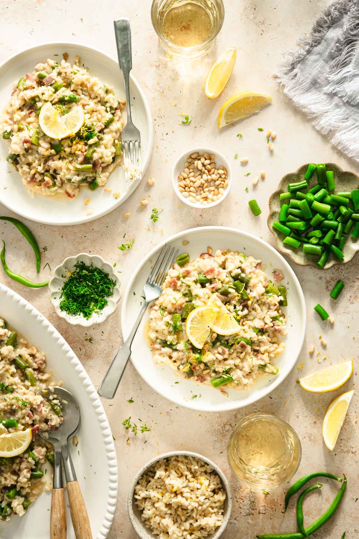 top view of green bean risotto on a serving plate, side of green beans, pine nuts, fresh dill, lemon wedges, glasses with wine