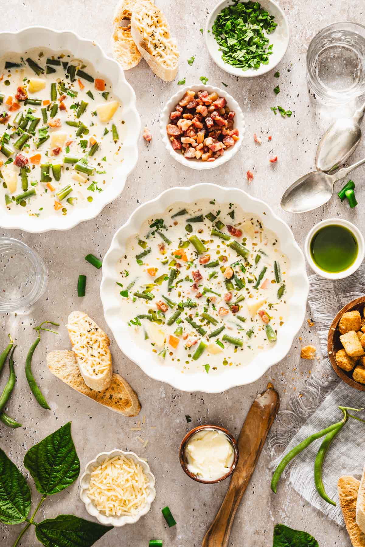 serving bowls with green bean soup, dish with bacon, green beans, glasses with water, dill oil, slices of bread