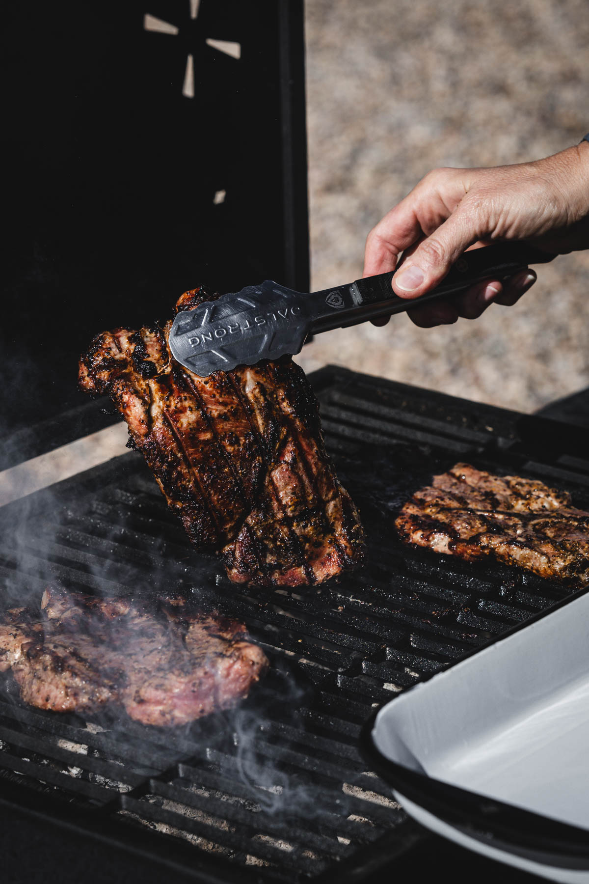 hands in frame holding tongs and piece of karkowka over grill grates
