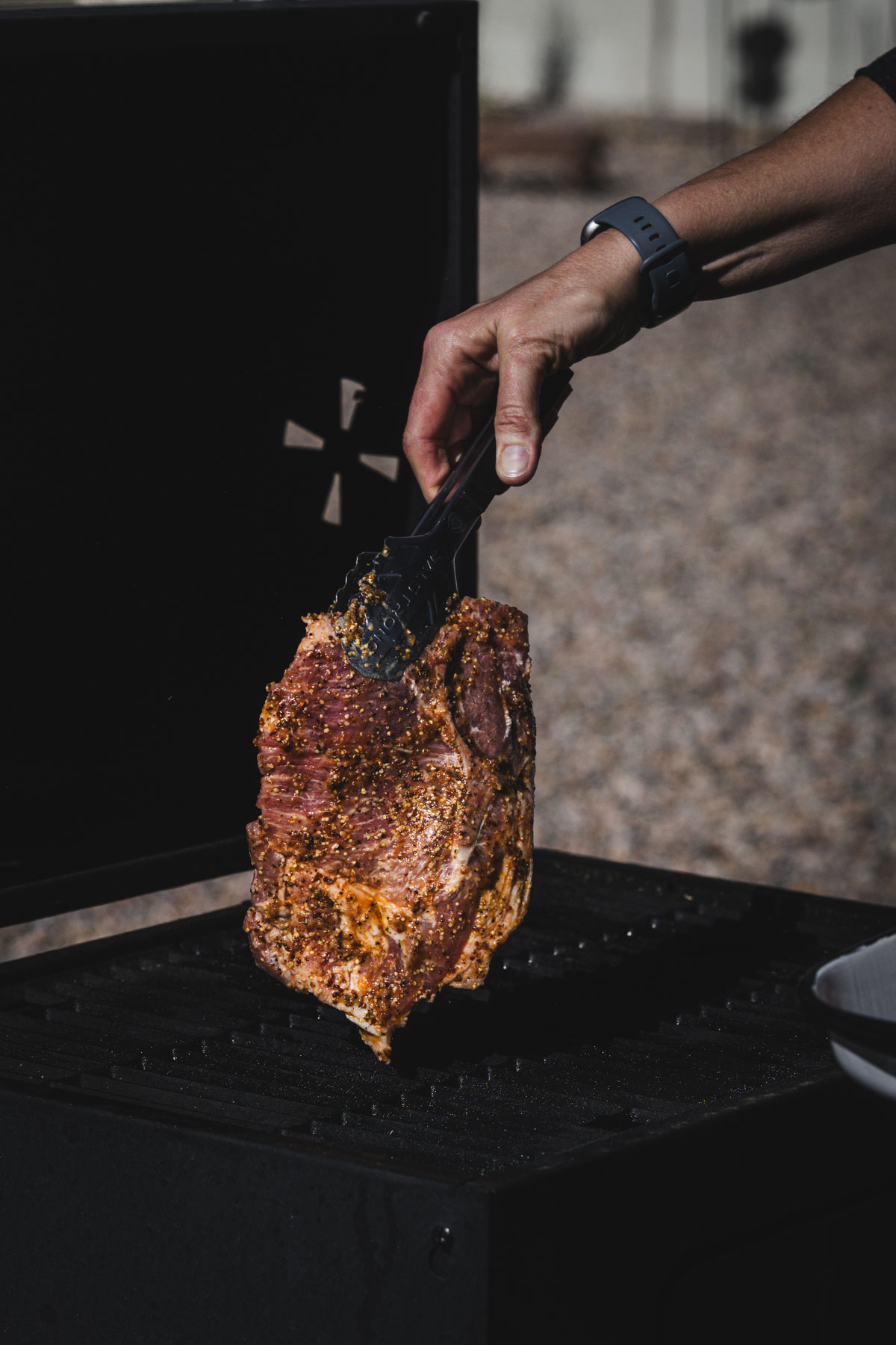 hands in frame holding tongs and piece of raw karkowka meat over grill grates