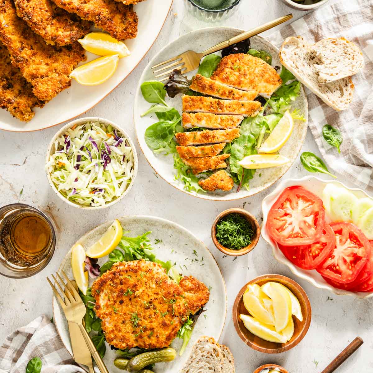 cooked kotlet presented on plates with salad, dish with fresh dill, glass of water, pickles, utensils and slices of bread, dish with salad and sliced tomatoes