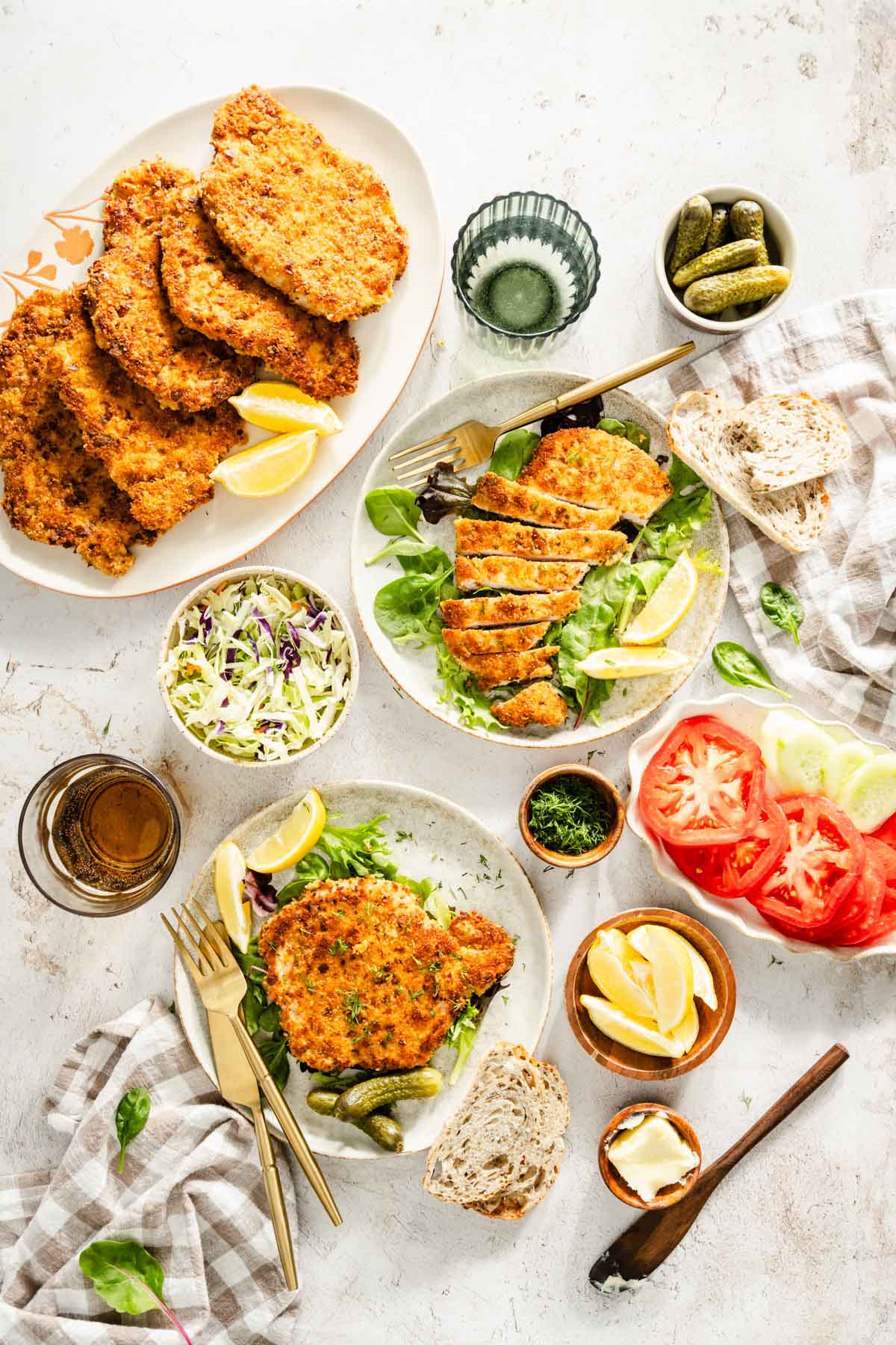 cooked por chops presented on plates with salad, dish with fresh dill, glass of water, pickles, utensils and slices of bread, dish with salad and sliced tomatoes