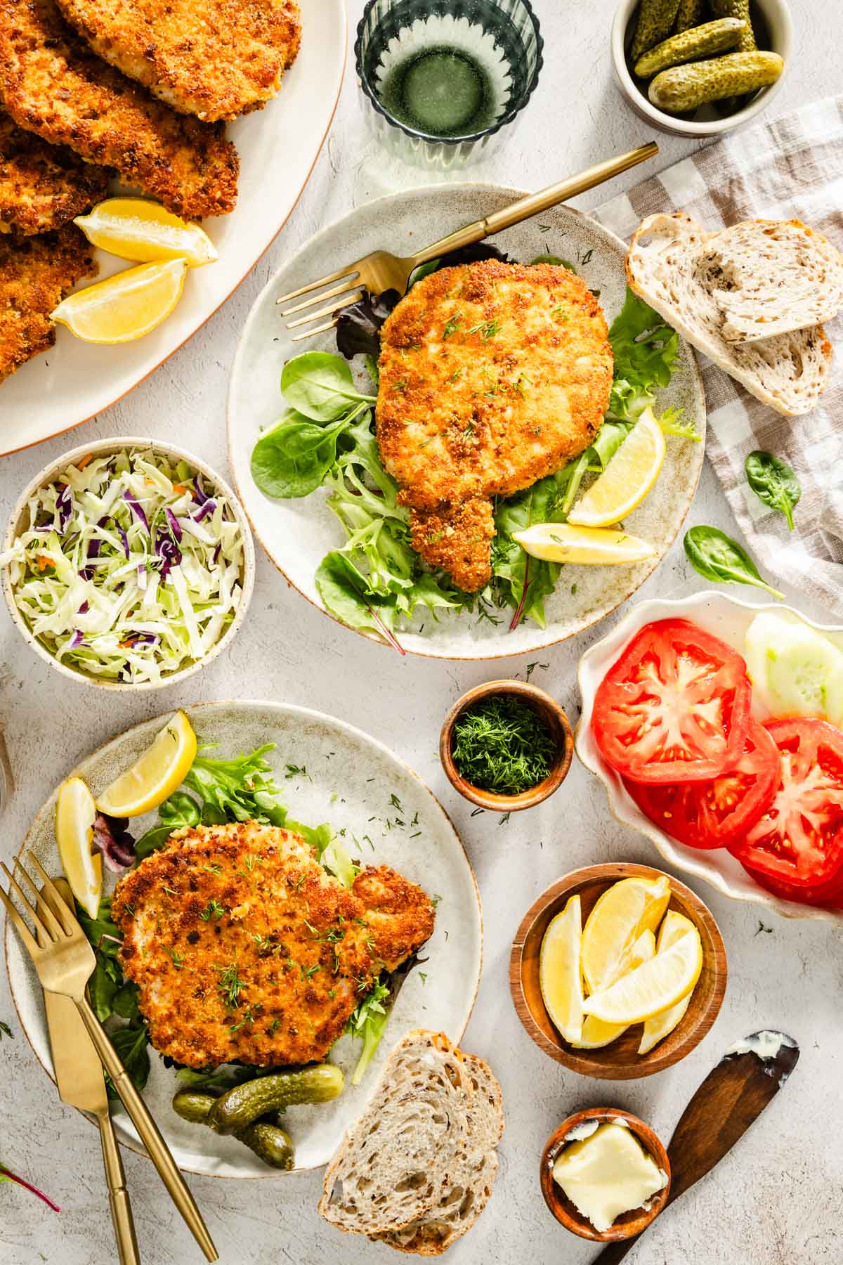 cooked por chops presented on plates with salad, dish with fresh dill, glass of water, pickles, utensils and slices of bread, dish with salad and sliced tomatoes