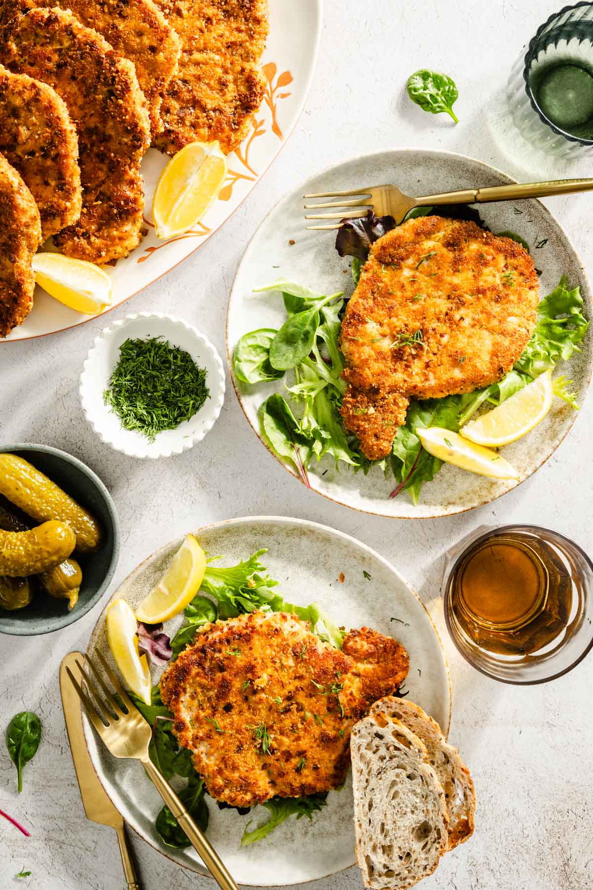 cooked por chops presented on plates with salad, dish with fresh dill, glass of water, pickles, utensils and slices of bread